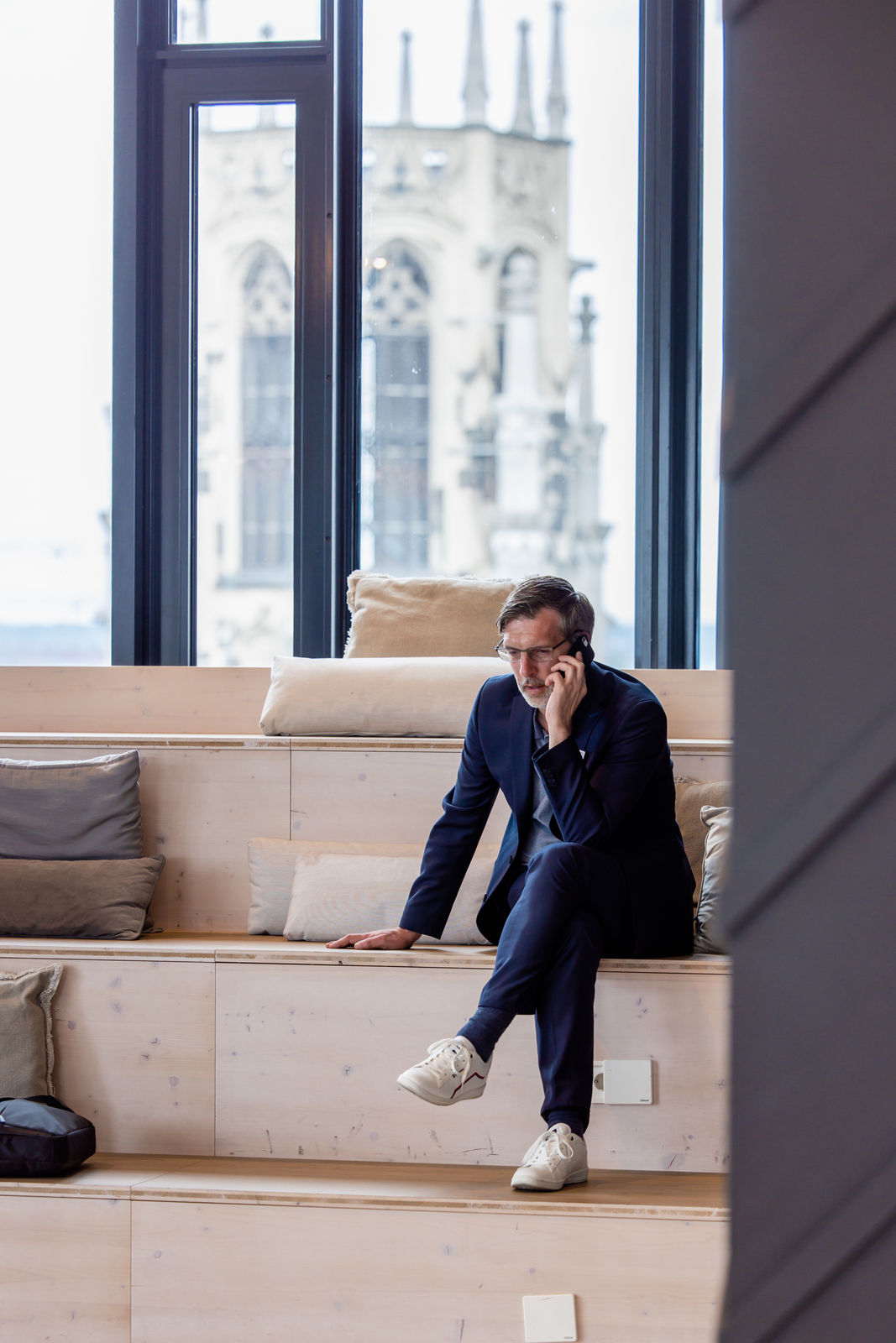 Man in dark suit sitting on wooden tiered seating, talking on a phone with cathedral windows in background.
