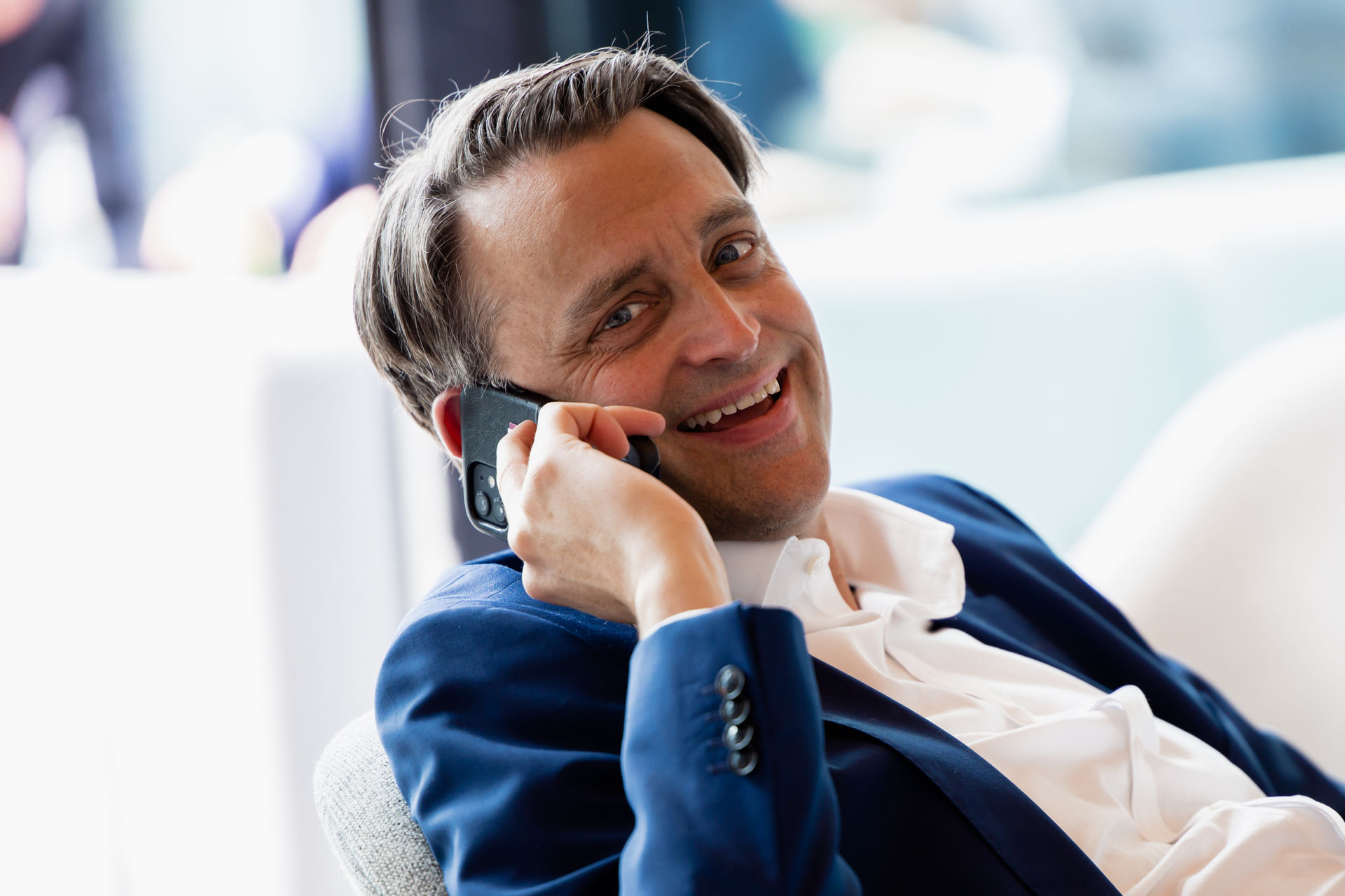 Smiling man in a blue suit talking on a smartphone while seated indoors.