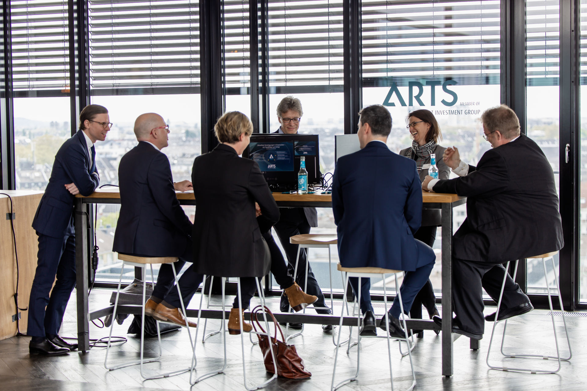 Six business professionals in formal attire having a discussion around a tall table with laptops and bottles inside an office with city view.