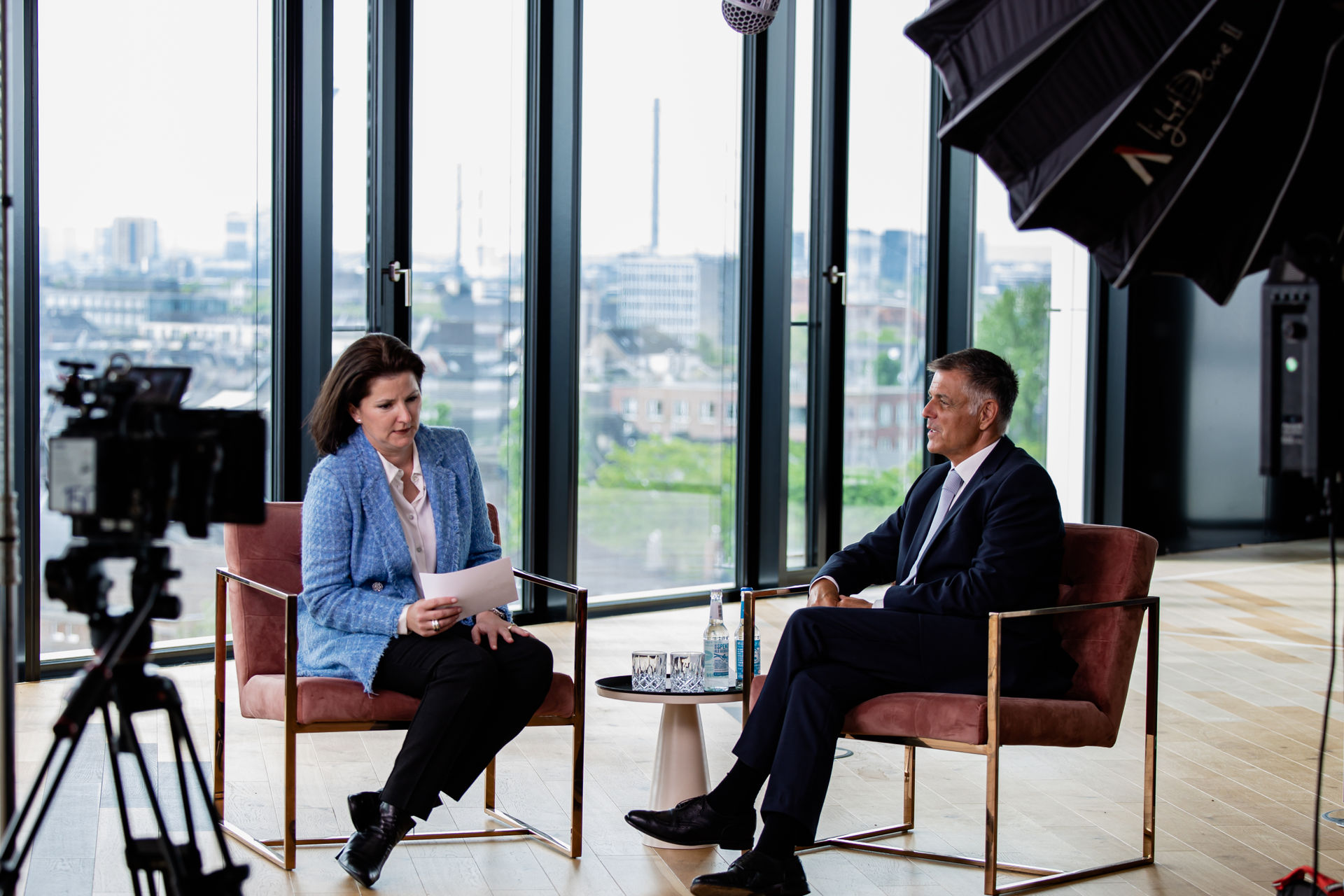 Two professionals seated facing each other in a modern office with large windows, recording an interview with camera and lighting equipment.