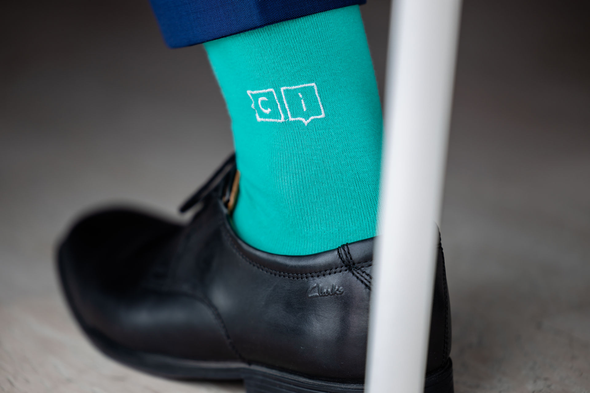 Close-up of a black leather dress shoe and bright teal sock with white outlined letters 'c' and 'i'.