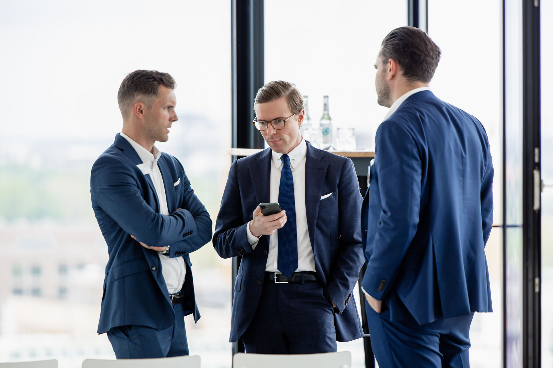 Three men in blue suits having a discussion in a bright office, one looking at his smartphone.