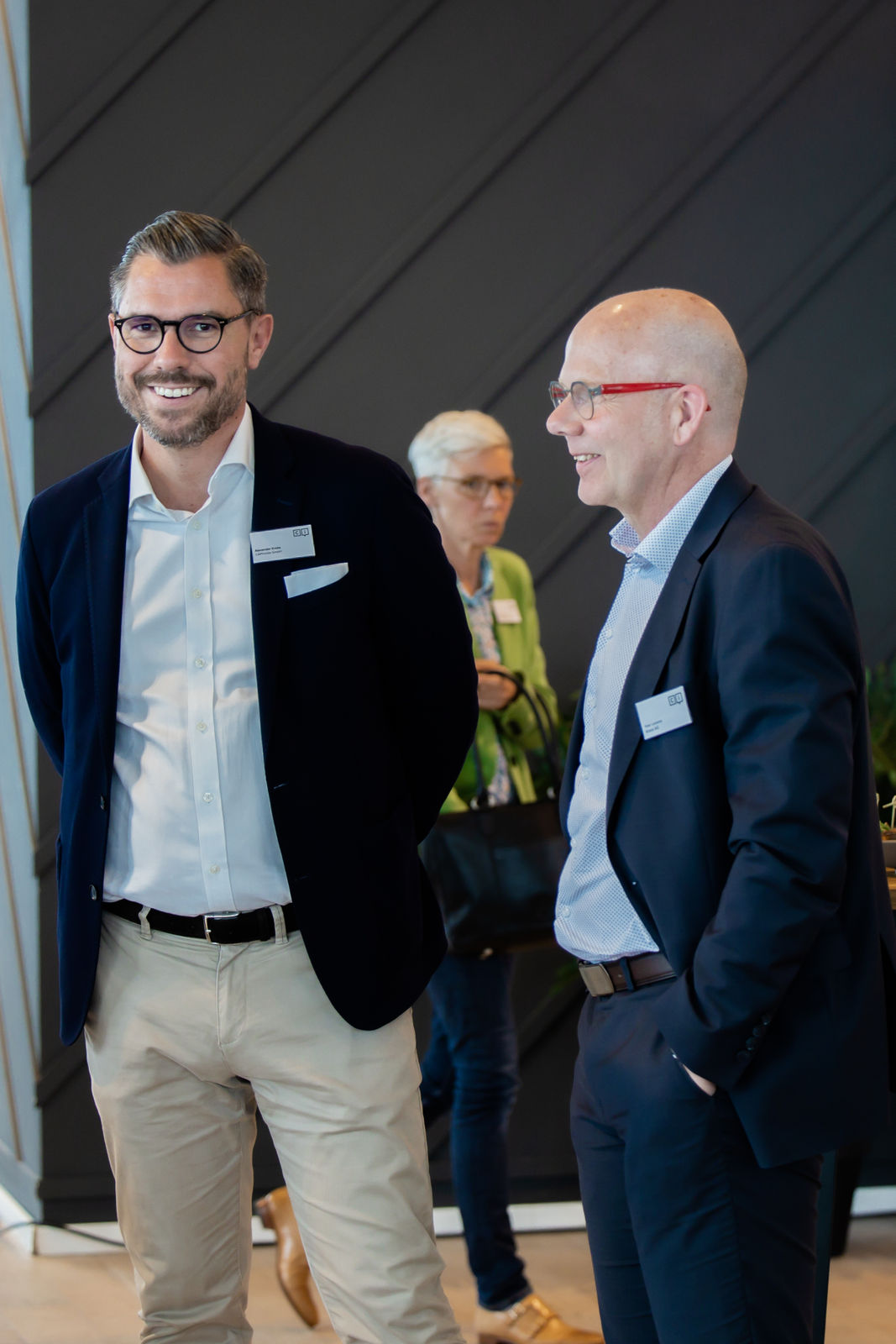 Two men in business attire smiling and conversing indoors, with a woman in a green jacket holding a black bag in the background.