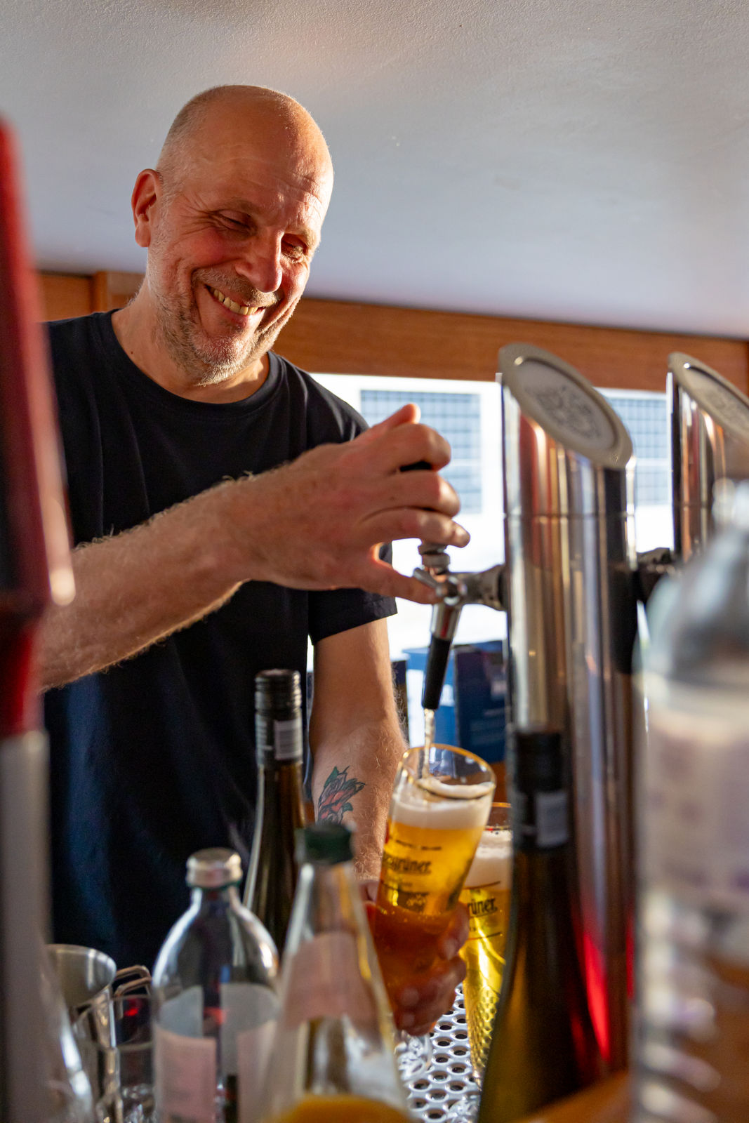 Smiling bartender pouring beer from a tap into a glass behind a bar with bottles in the foreground.