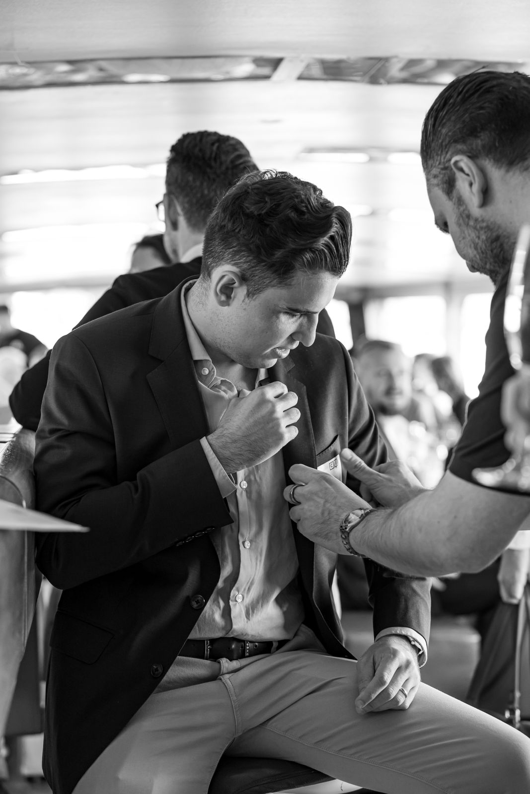 Man seated adjusting or pinning a name tag on his blazer with another man assisting him in a crowded indoor setting.
