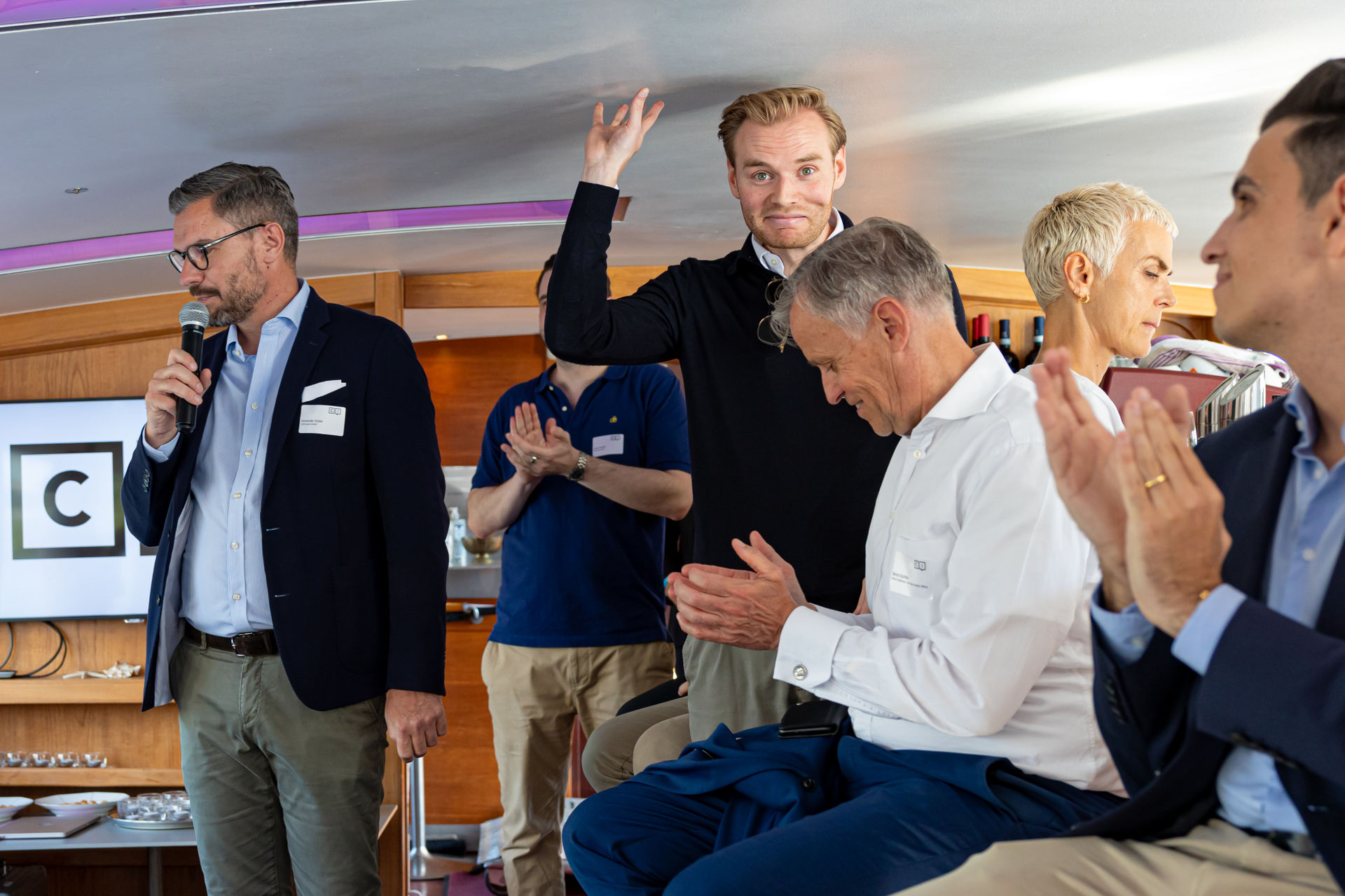 Man in blazer speaking into microphone while others around him applaud and one man raises his hand in acknowledgment in a conference room setting.