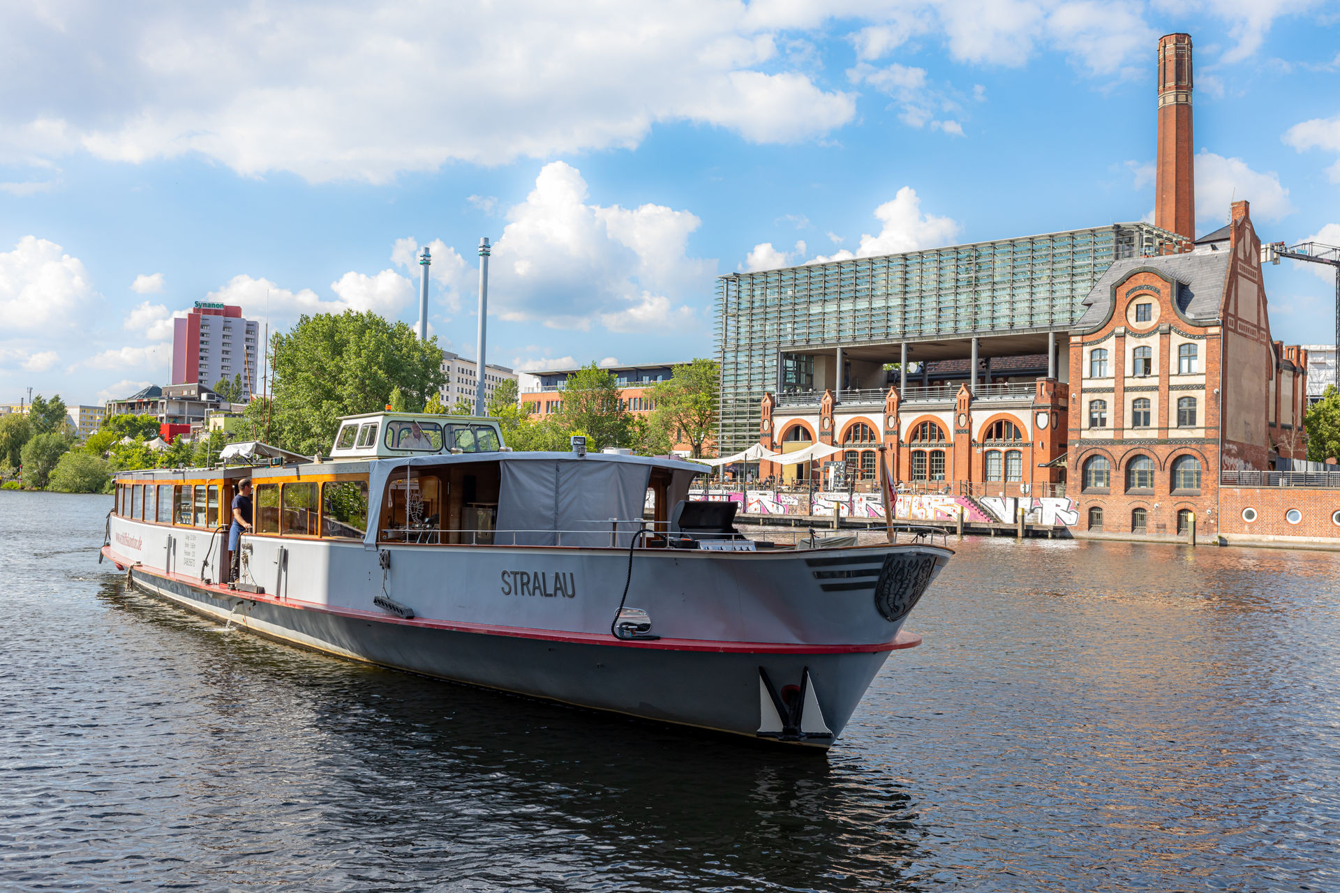 Gray passenger boat named STRALAU sailing on a river with modern and historic buildings in the background under a partly cloudy sky.