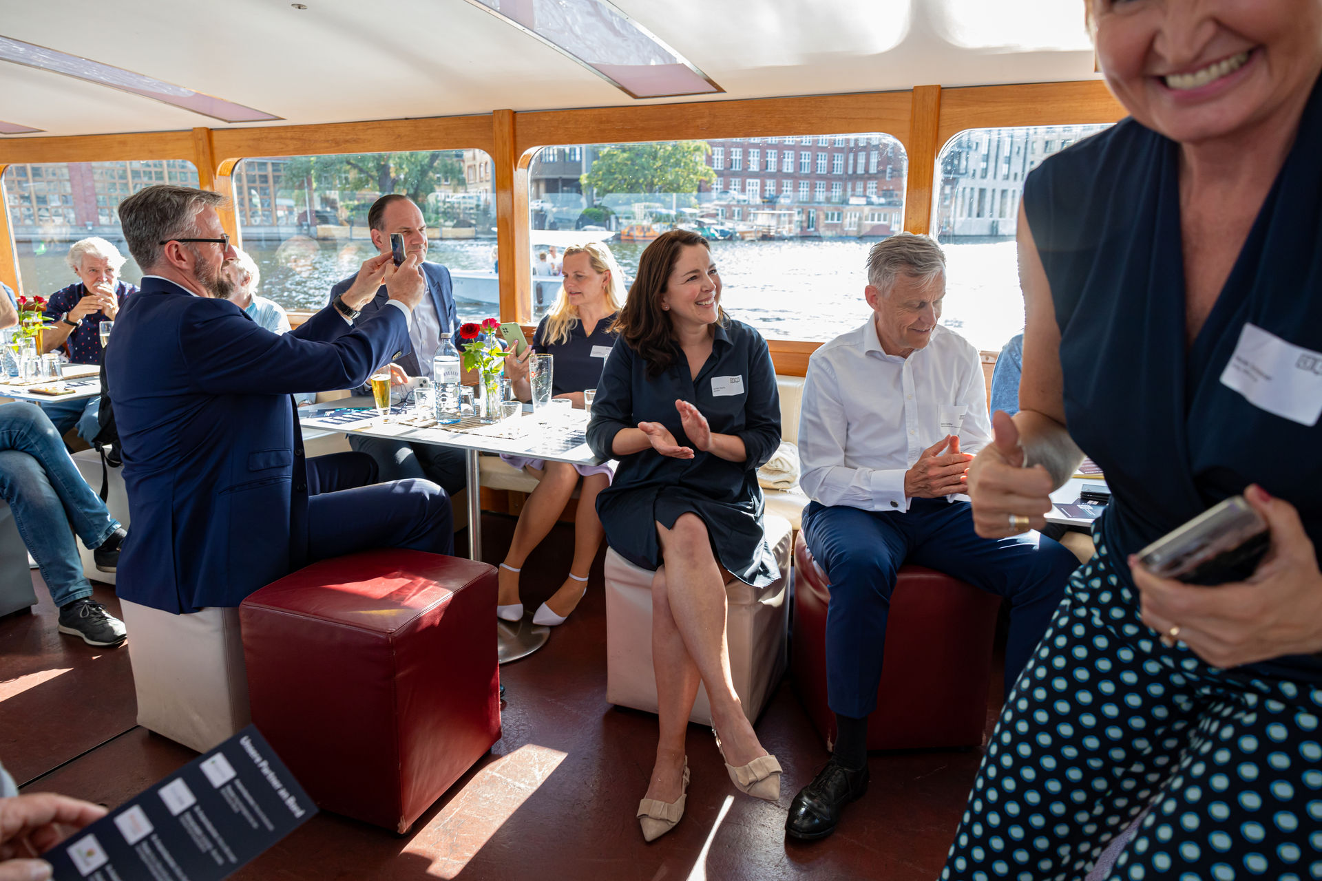Group of people socializing inside a boat with large windows showing water and buildings outside, some clapping and smiling.