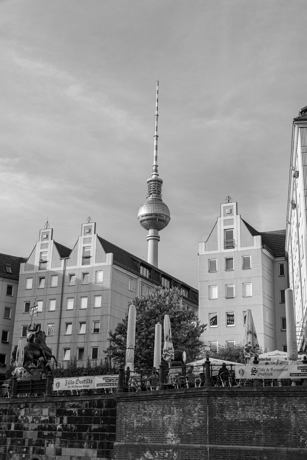 View of Berlin TV Tower rising behind traditional buildings and an outdoor cafe with umbrellas and a statue in front.