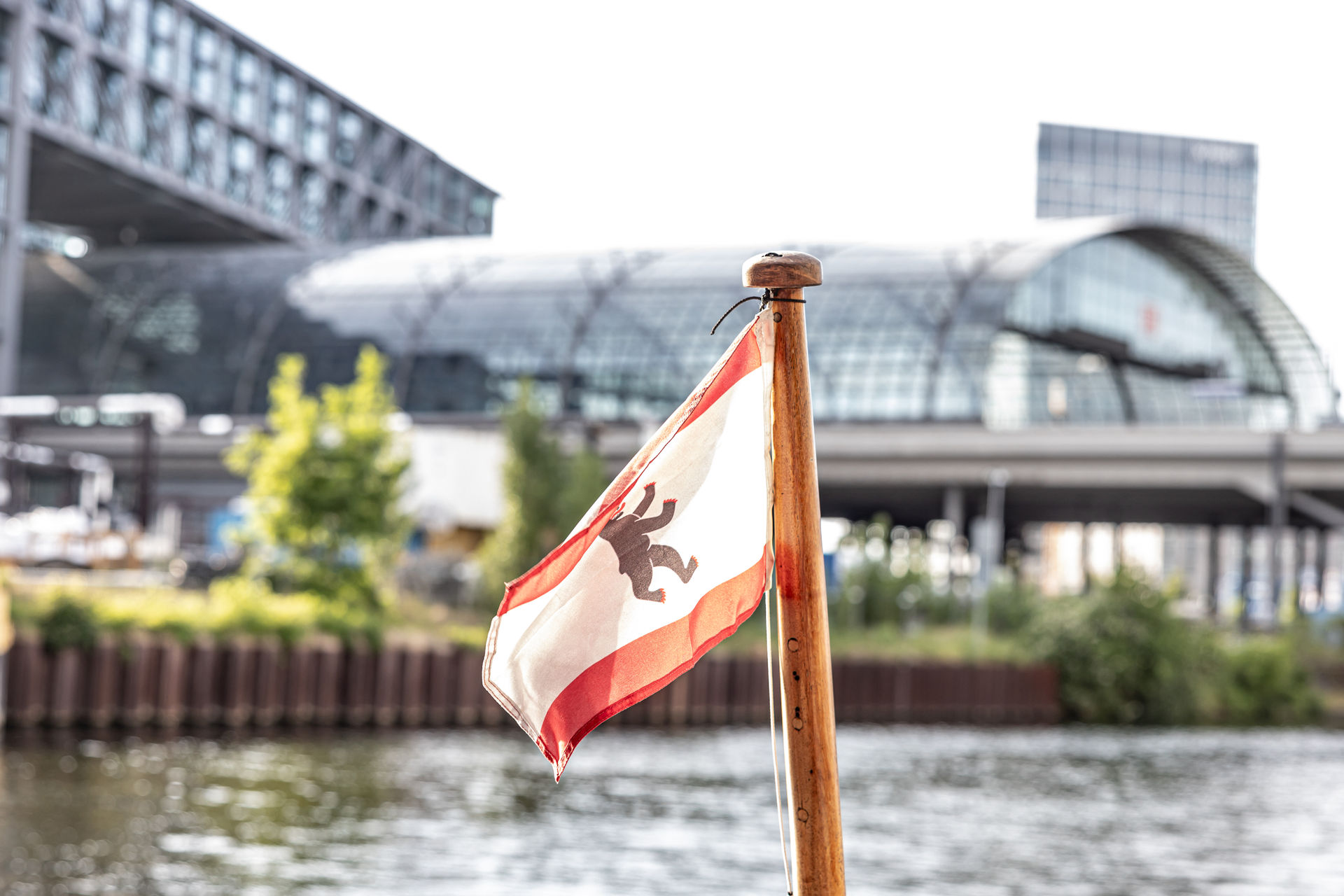 Small flag with a bear emblem on a wooden pole near water, with a modern glass building in the background.