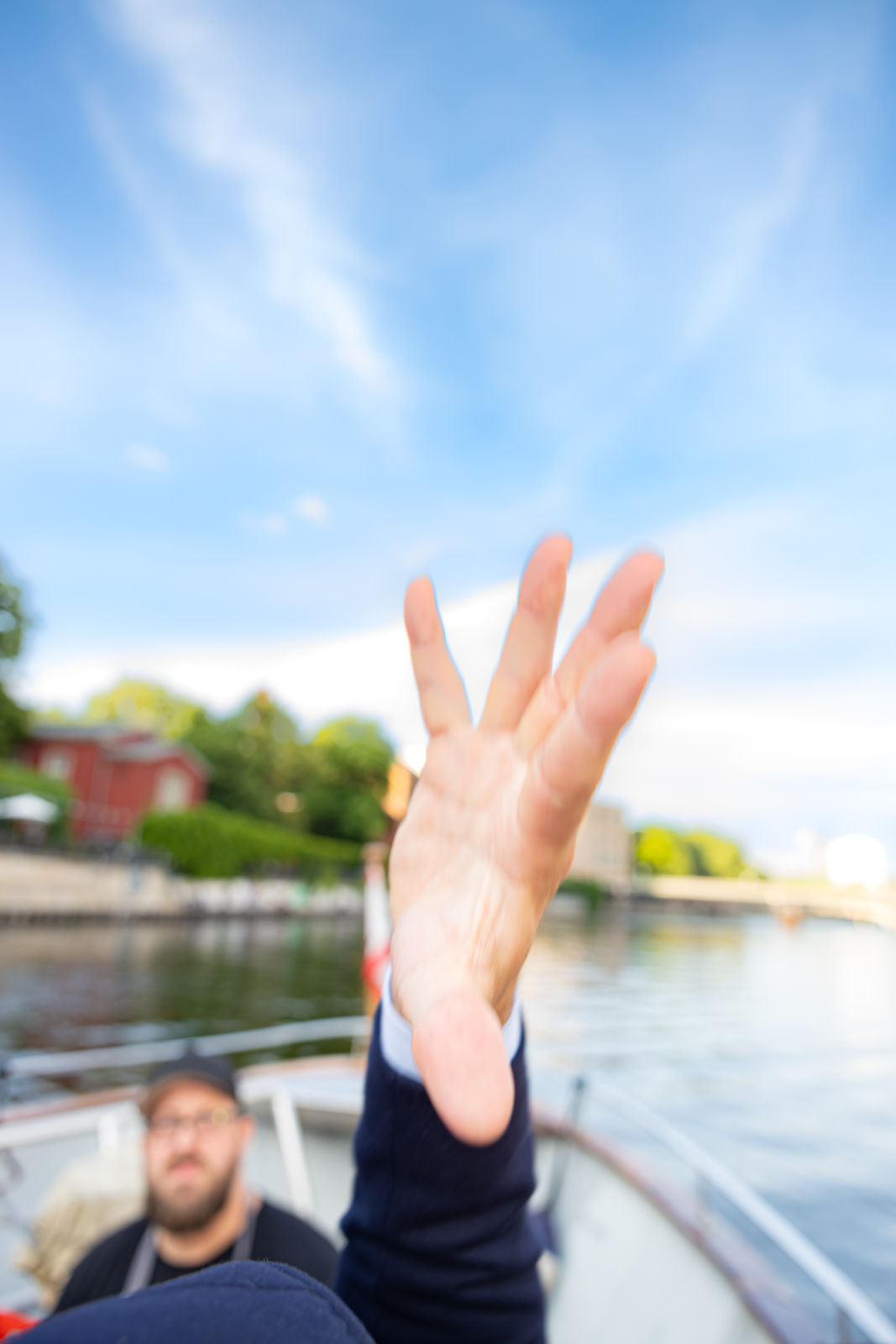 Close-up of a person’s hand reaching towards the camera on a boat with a blurred background of water, trees, and buildings.