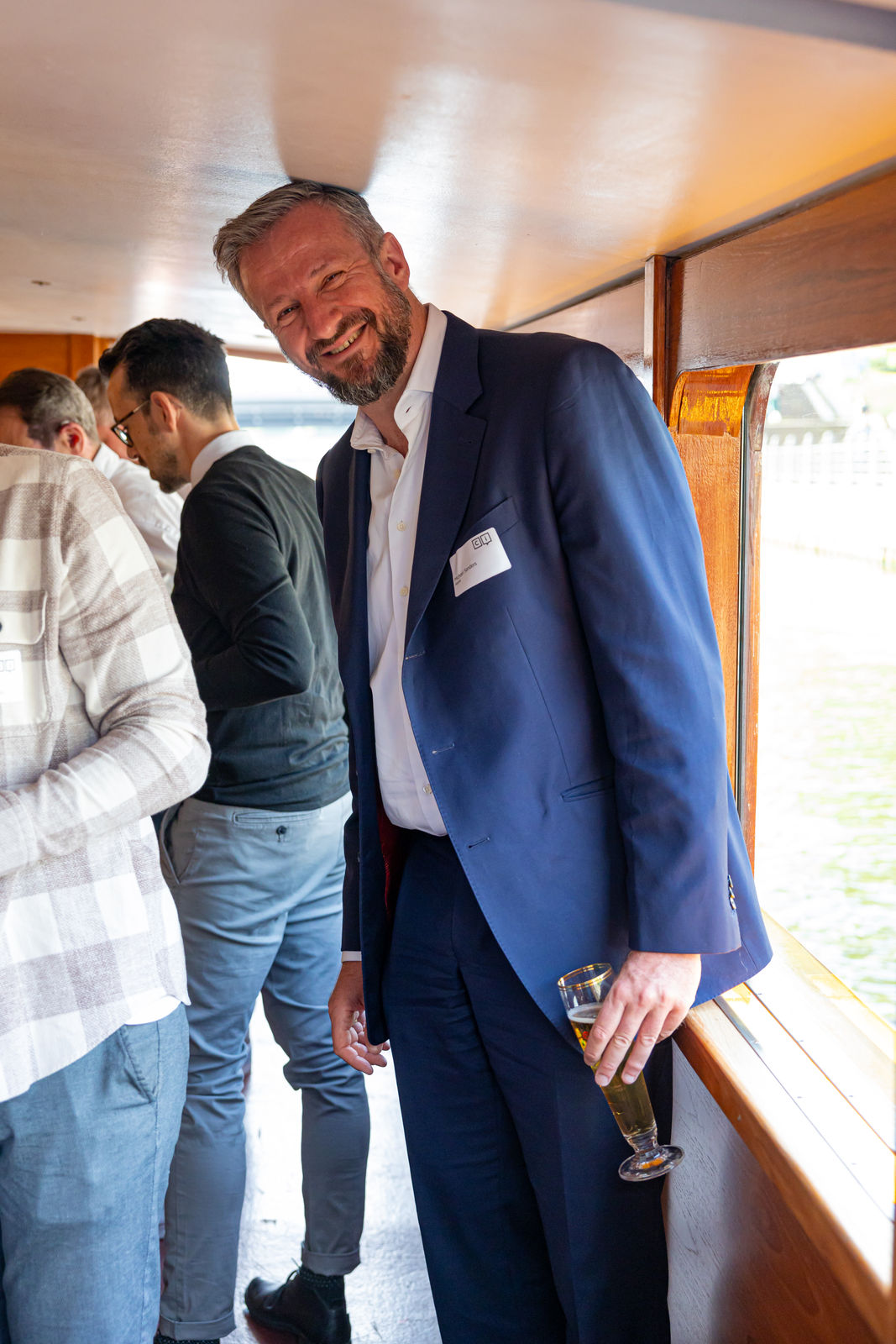 Smiling man in a navy blue suit holding a glass of beer, leaning on the wooden interior wall of a boat with other people in the background.