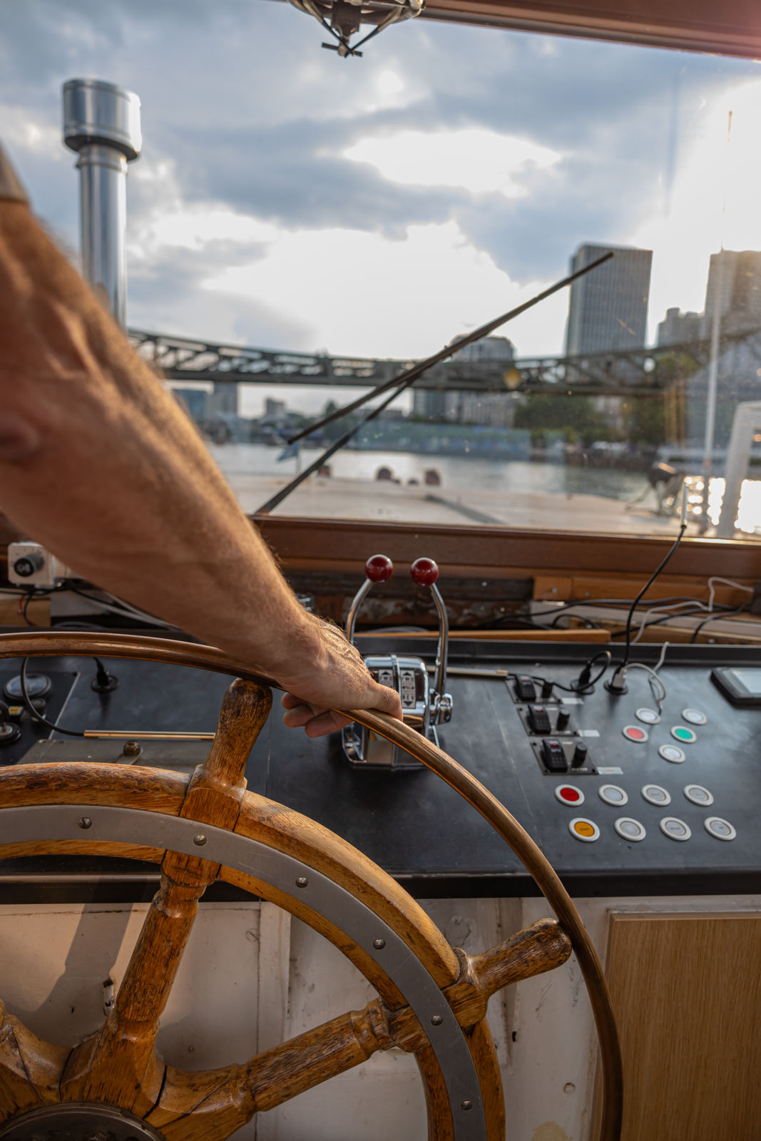 Person steering a wooden ship's wheel at a boat control panel with river and city buildings in the background.