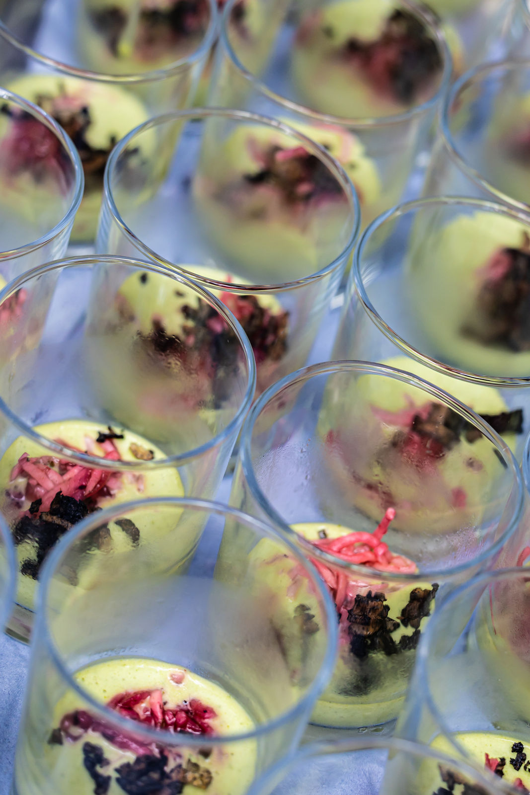 Close-up of multiple clear glasses containing yellow creamy dessert topped with pink and brown garnishes.