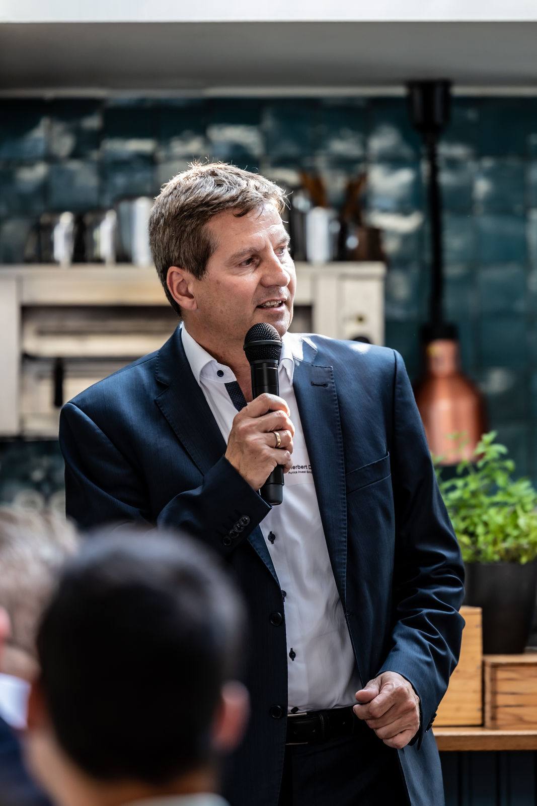 Man in a navy suit speaking into a microphone during an indoor event.