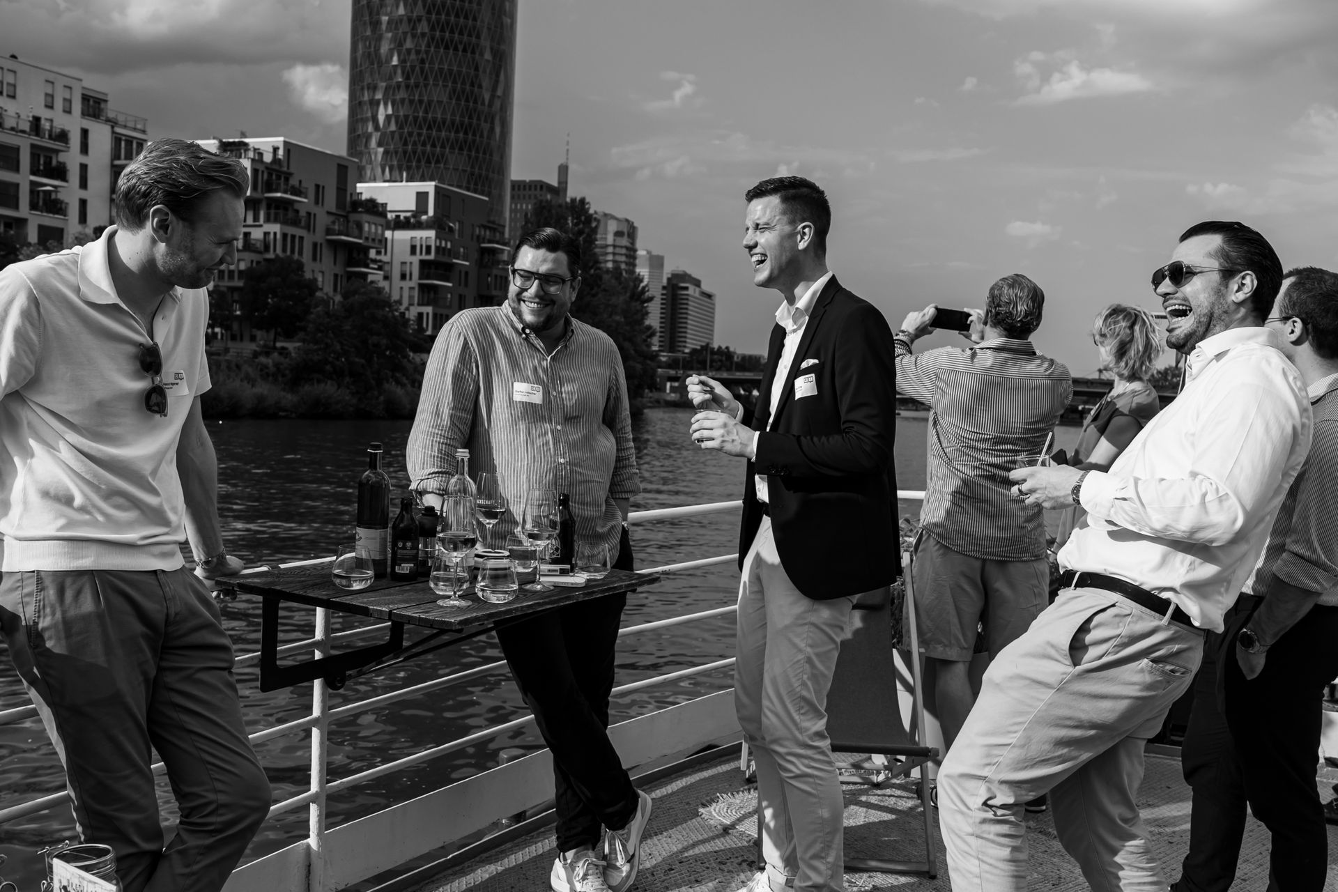 Group of men laughing and socializing on a boat deck near a river with city buildings in the background.