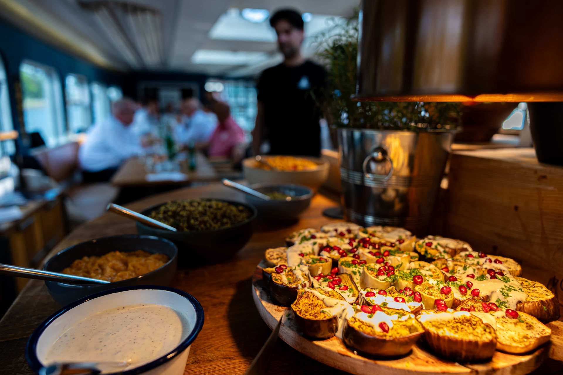 Wooden table with a platter of roasted eggplant slices topped with sauce and pomegranate seeds, alongside several bowls of sauces and dips.