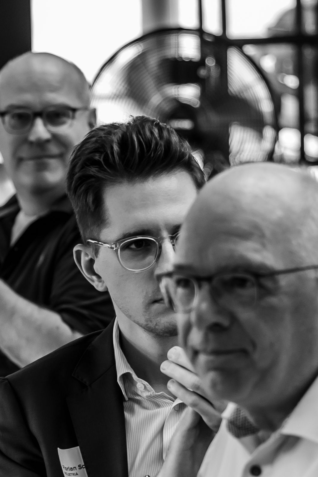 Black and white photo of three men wearing glasses, with the youngest man in focus and the other two older men blurred in the foreground and background.