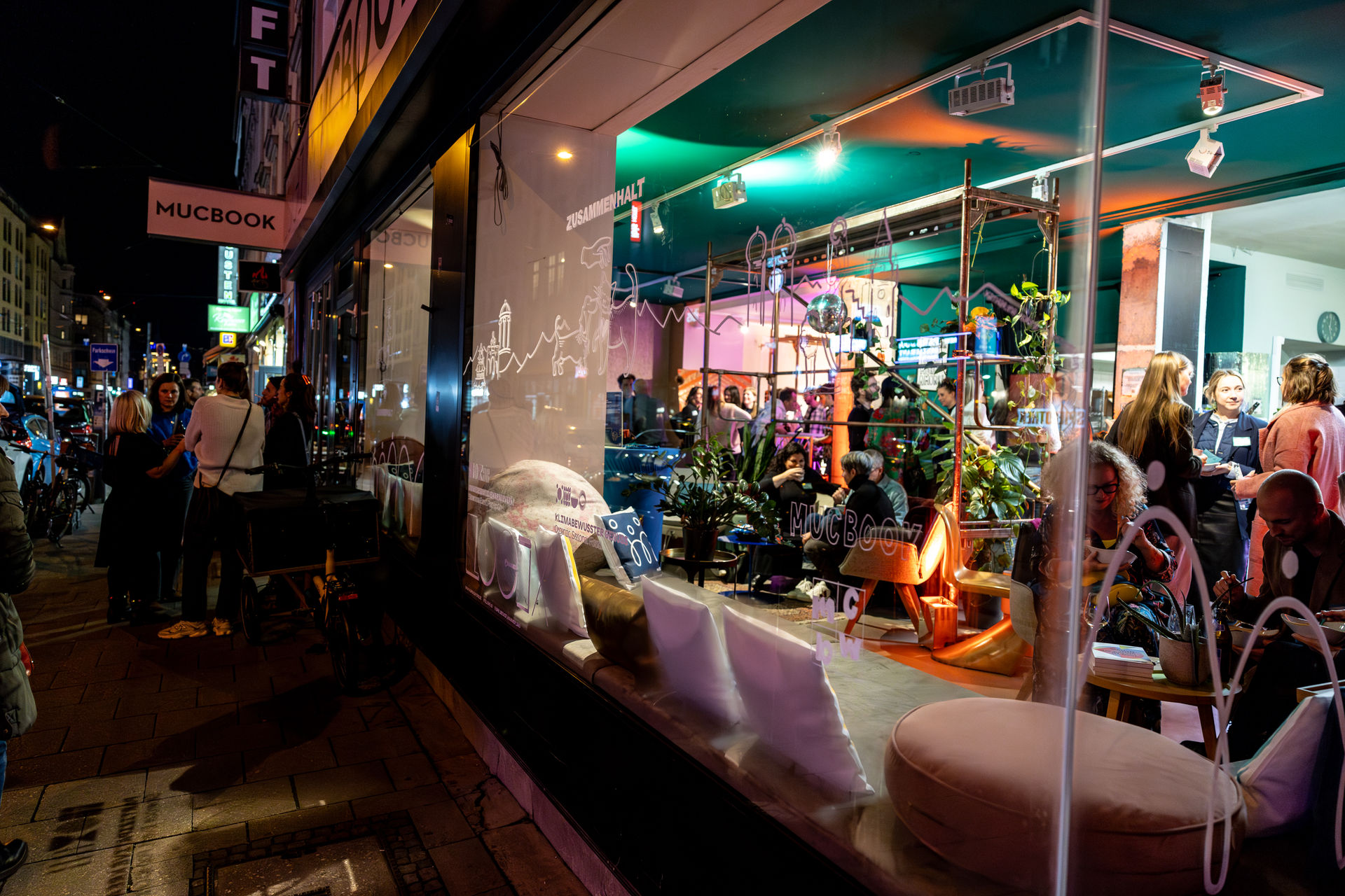 Nighttime street view of a lively café or bookstore named MUCBOOK with people socializing inside and outside.