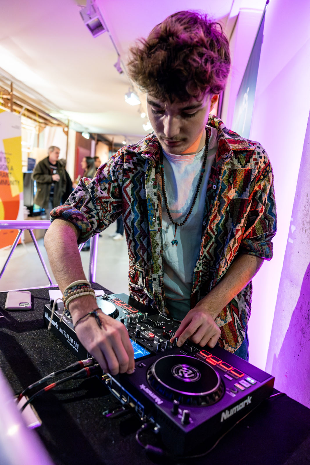 Young man wearing a colorful patterned shirt DJing with a Numark Mixstream Pro controller under pink and purple lighting.