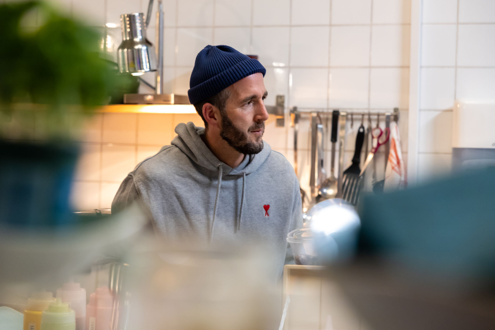Man wearing a blue knit beanie and gray hoodie looking to the side in a kitchen with utensils on the tiled wall behind him.