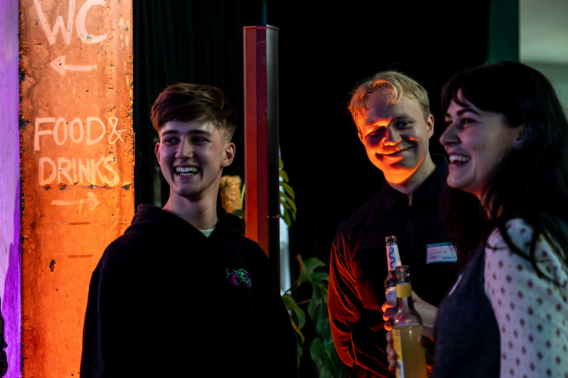 Three young people smiling and holding drinks at an indoor social gathering with a lit wall sign indicating 'WC' and 'Food & Drinks'.