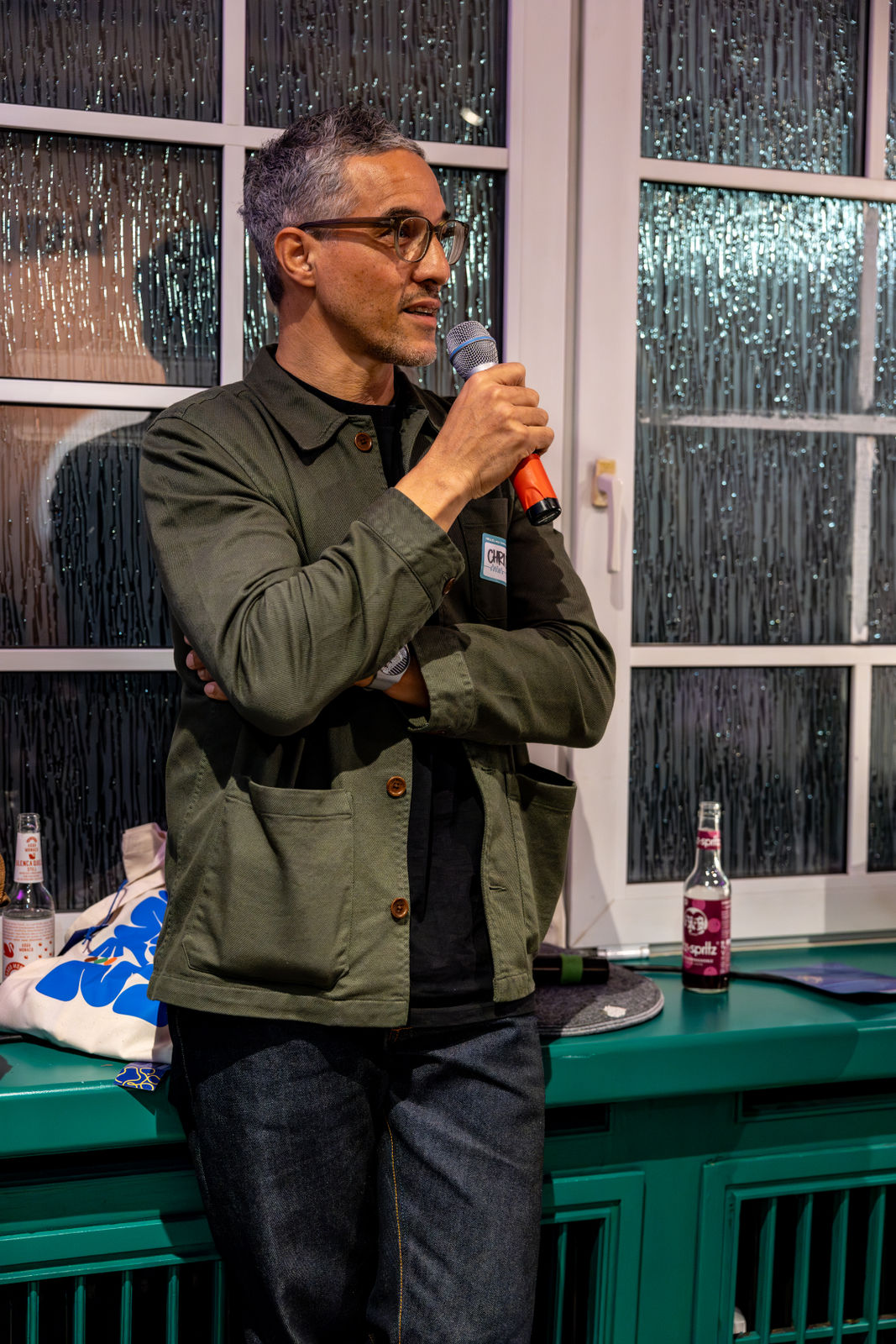 Man with glasses and gray hair speaking into a microphone indoors by a window with textured glass.