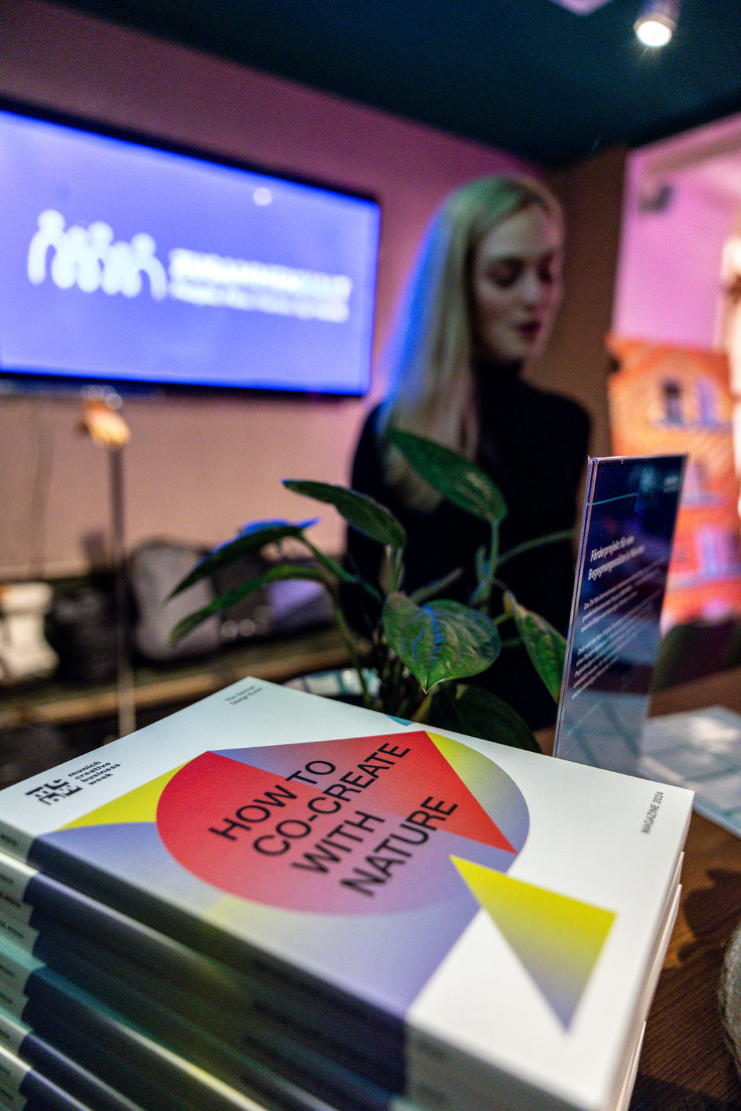 Stack of books titled 'How to Co-Create with Nature' on a table with a green plant and a blurred person in the background.