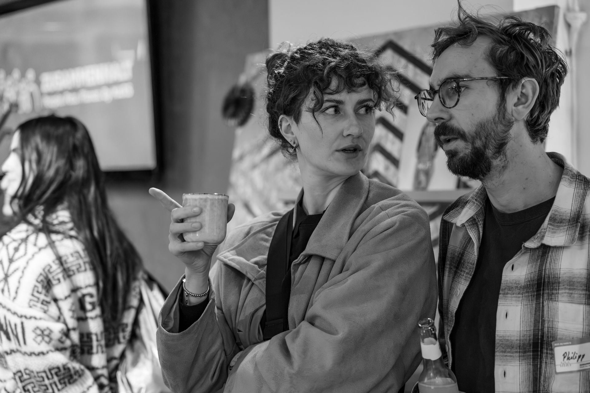 Black and white photo of a woman holding a cup and pointing while talking to a man with glasses holding a bottle.