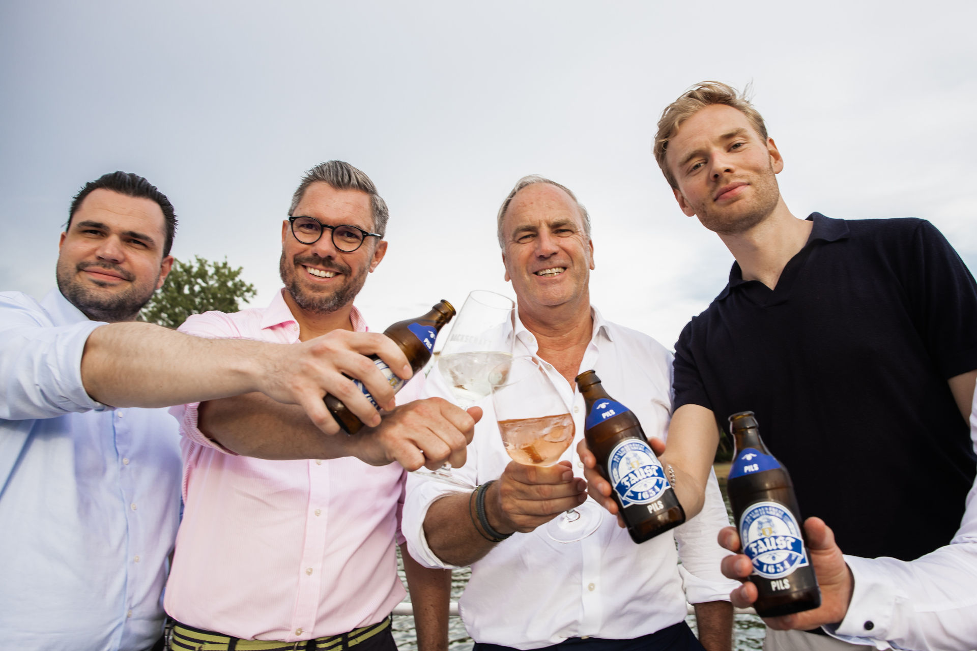 Four men smiling and toasting with beer bottles and wine glasses outdoors near water.