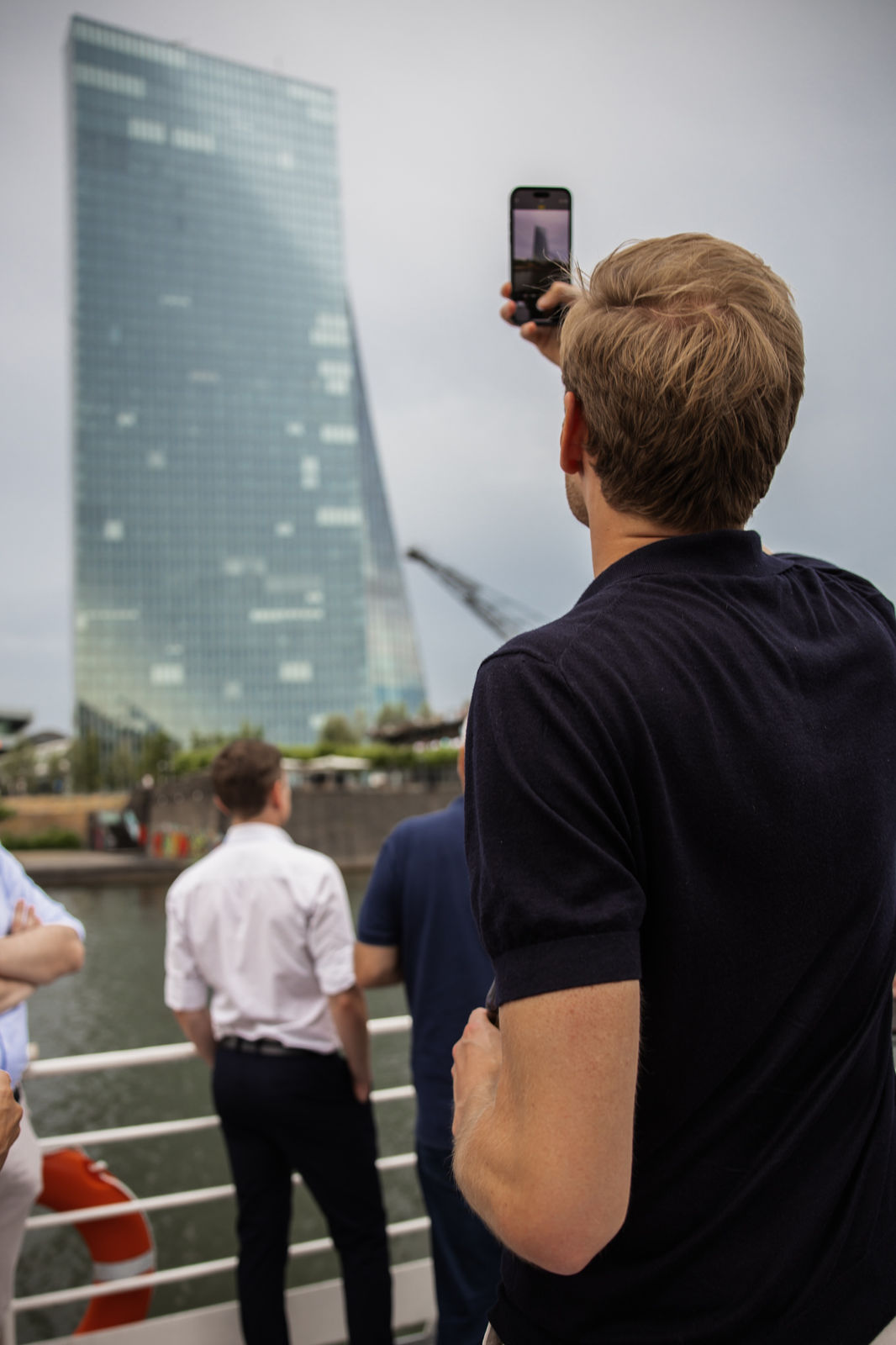 Man with blonde hair taking a photo of a tall glass building by the water from a boat deck with other people.