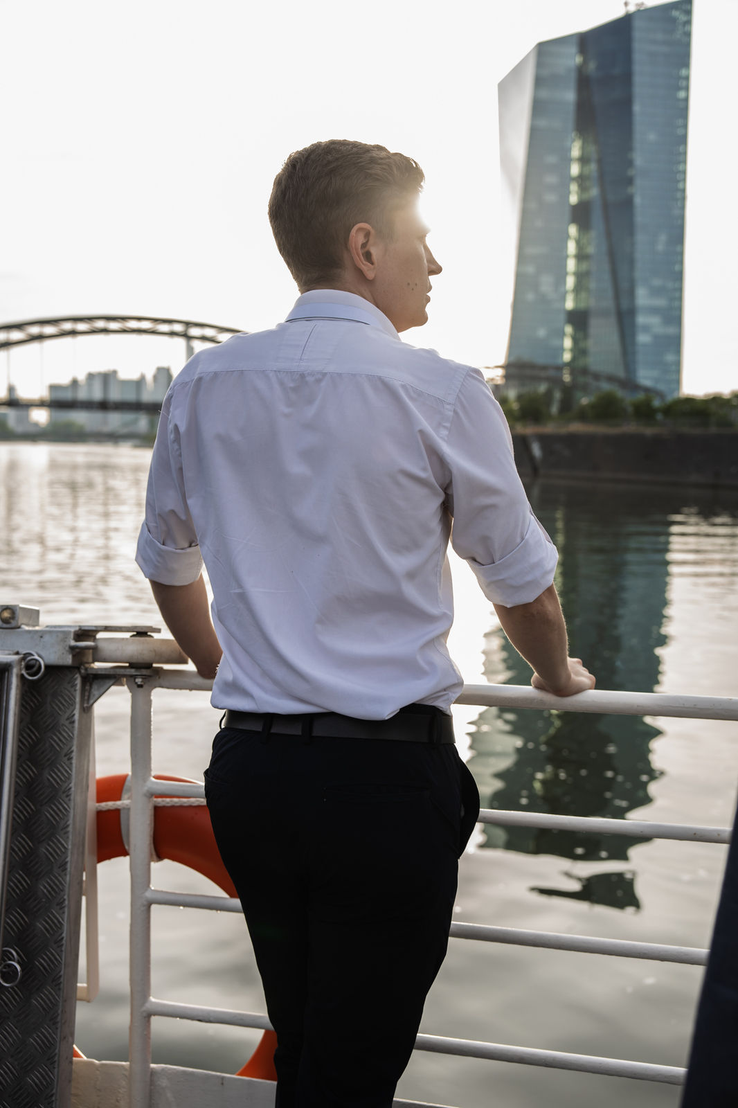 Man in white shirt and black pants standing on a boat railing, looking at a modern glass building across the water.