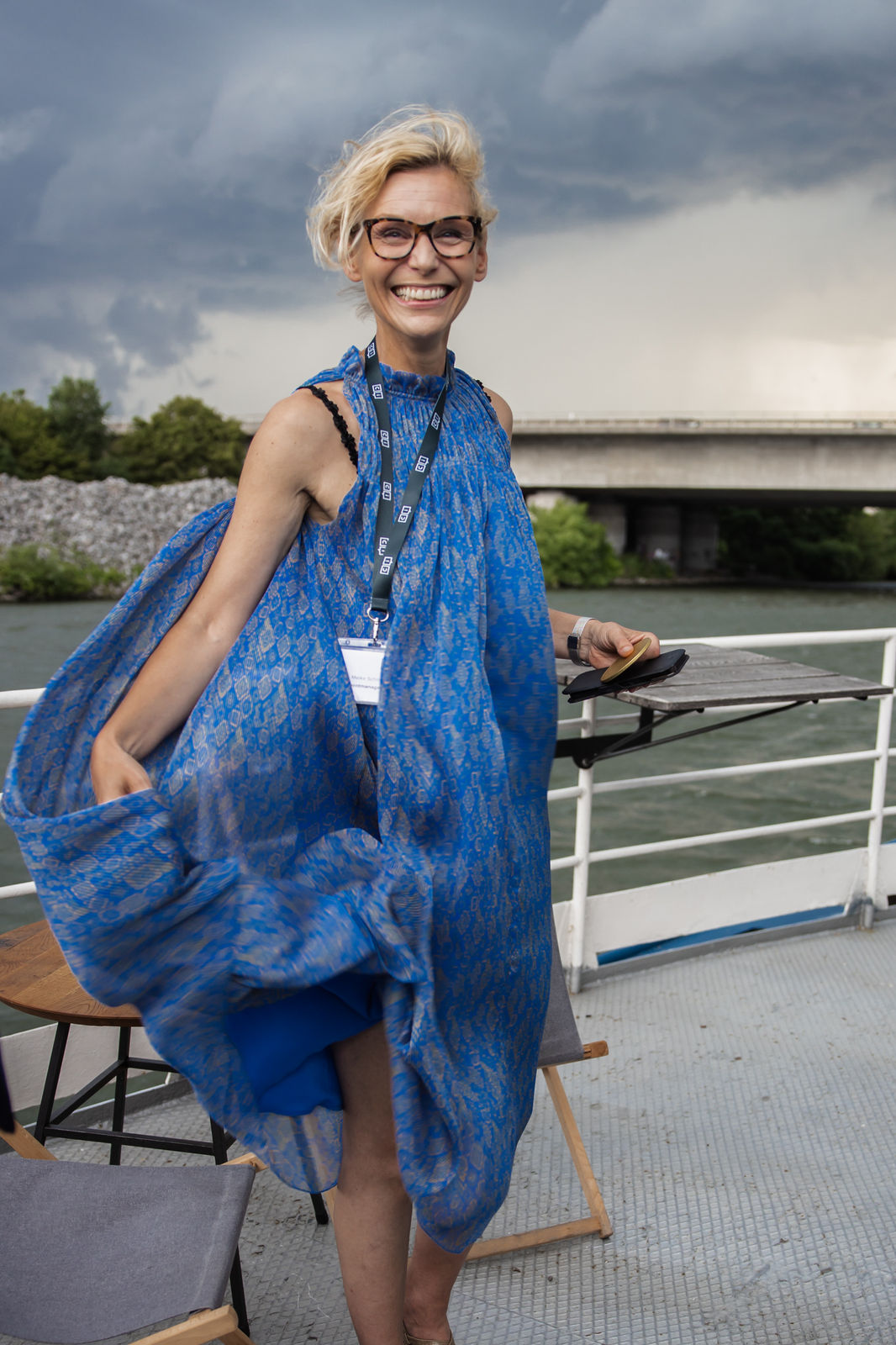 Smiling woman with short blonde hair wearing glasses and a flowing blue dress on a boat deck with river and bridge in the background.