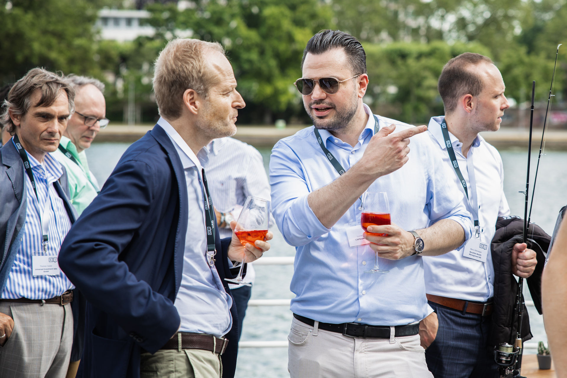 Five men in casual business attire socializing outdoors near water, two holding glasses of orange drink, one holding fishing rods.