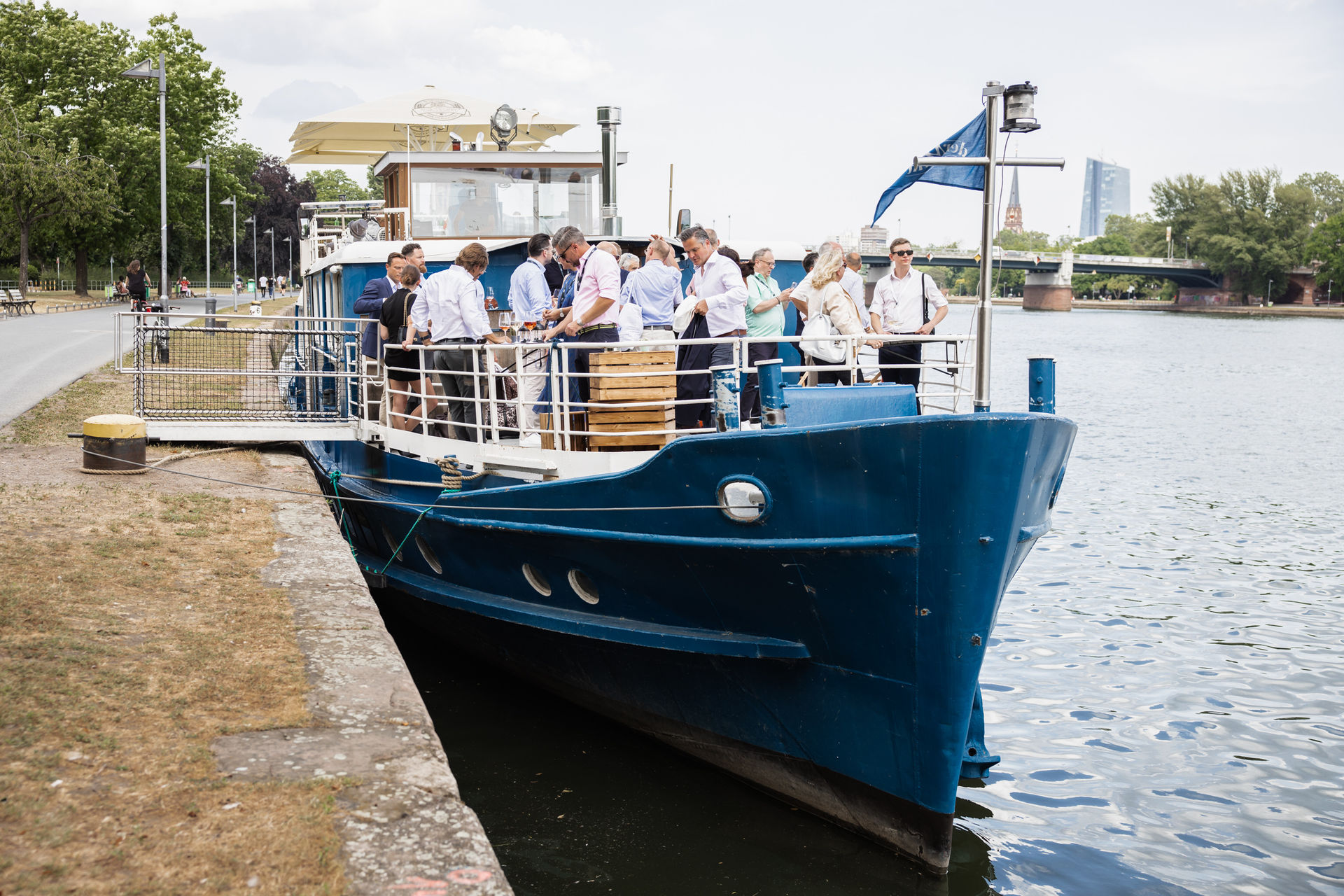 Group of people dressed in semi-formal attire gathered on the deck of a blue boat docked along a riverside with a city skyline in the background.