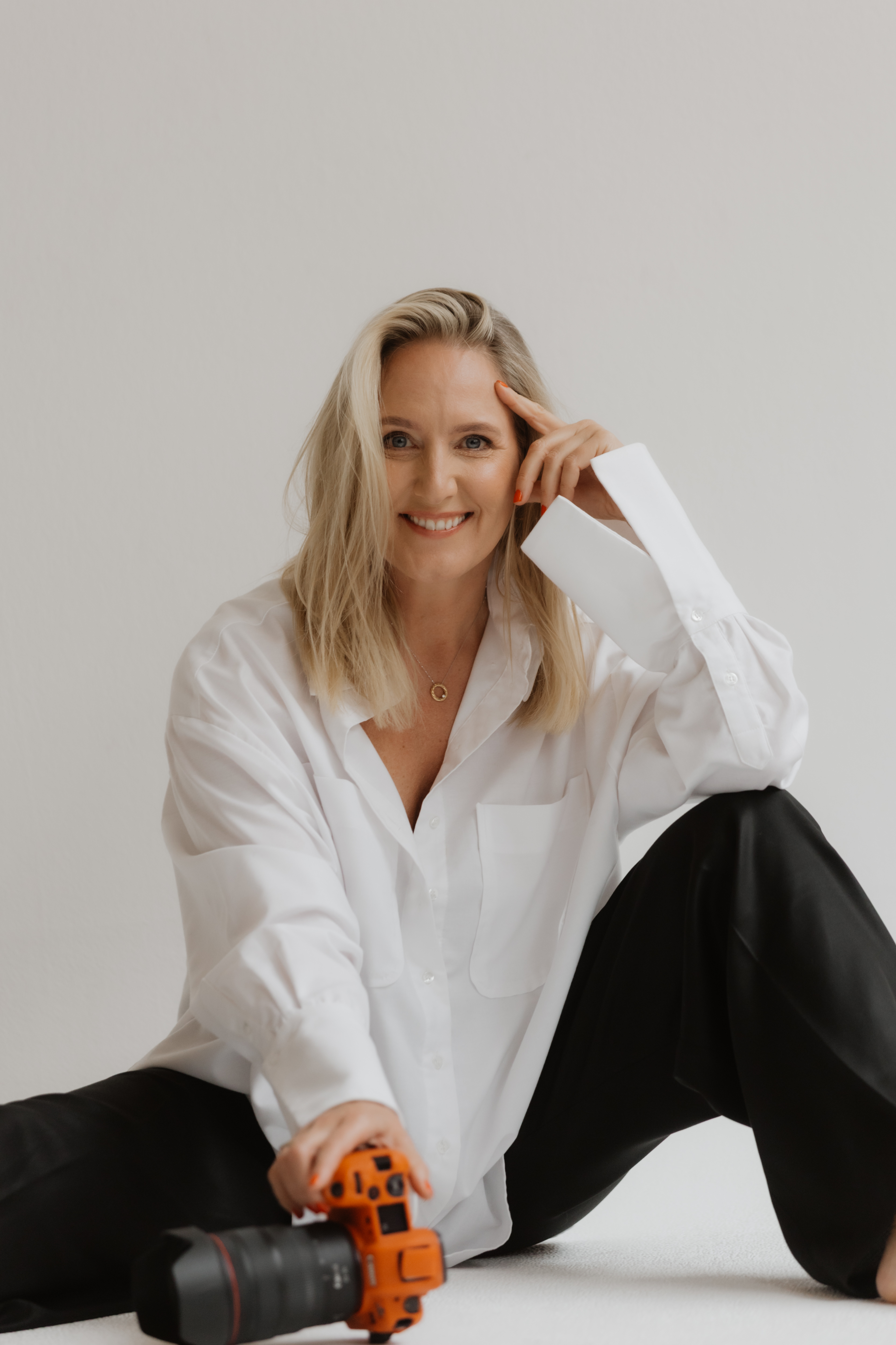 Smiling woman with blonde hair sitting on floor wearing a white shirt and black pants, holding an orange camera.
