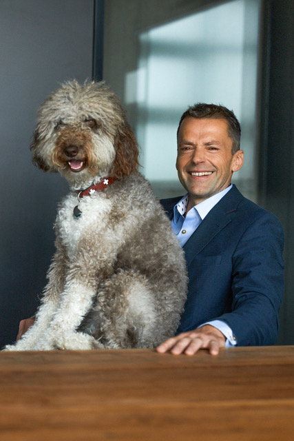 Man in a blue suit smiling with a large curly-haired dog sitting on a table in front of him.