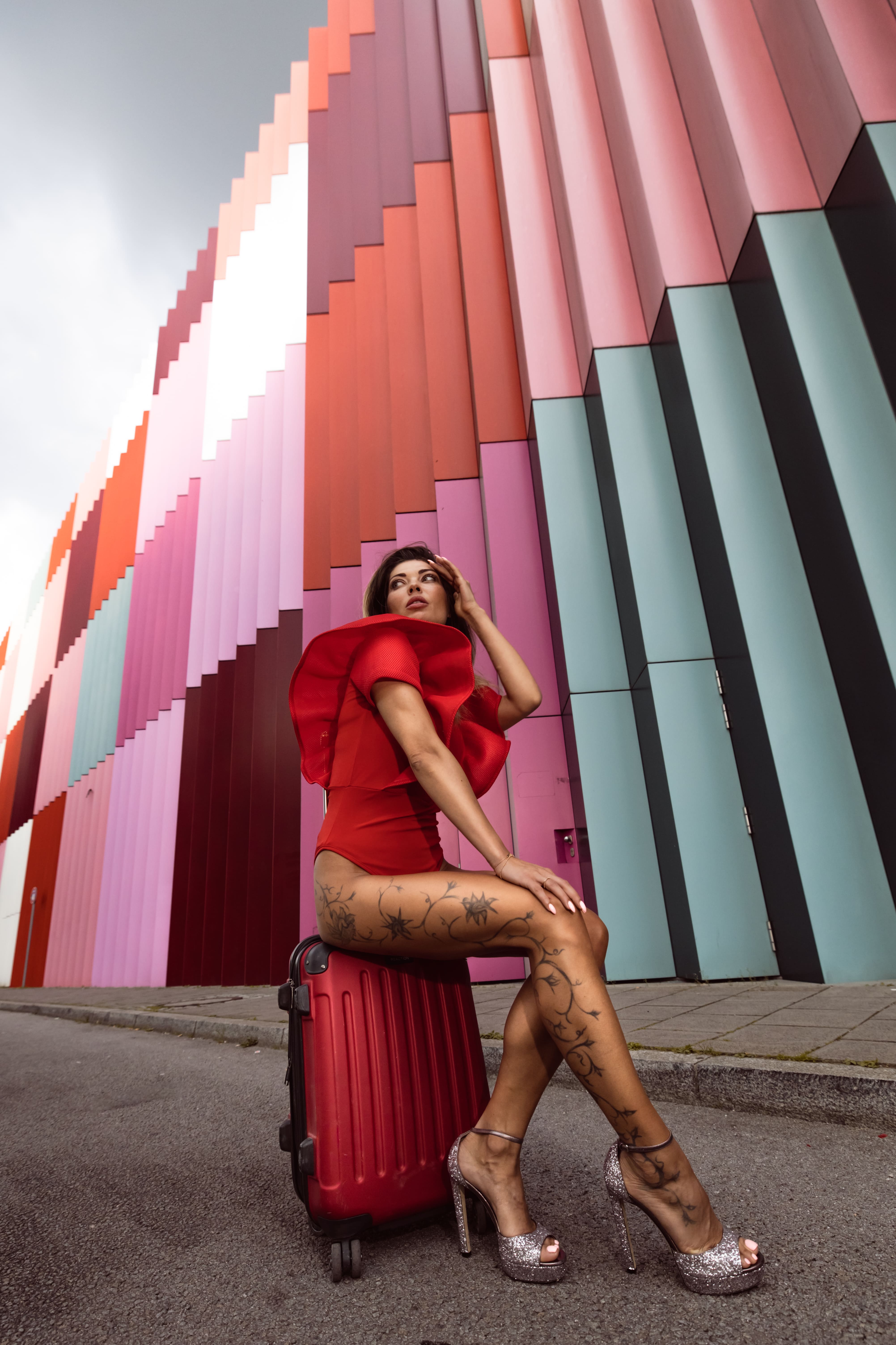 Woman in a red outfit and glittery high heels sitting on a red suitcase with floral tattoos on her legs against a colorful urban wall.