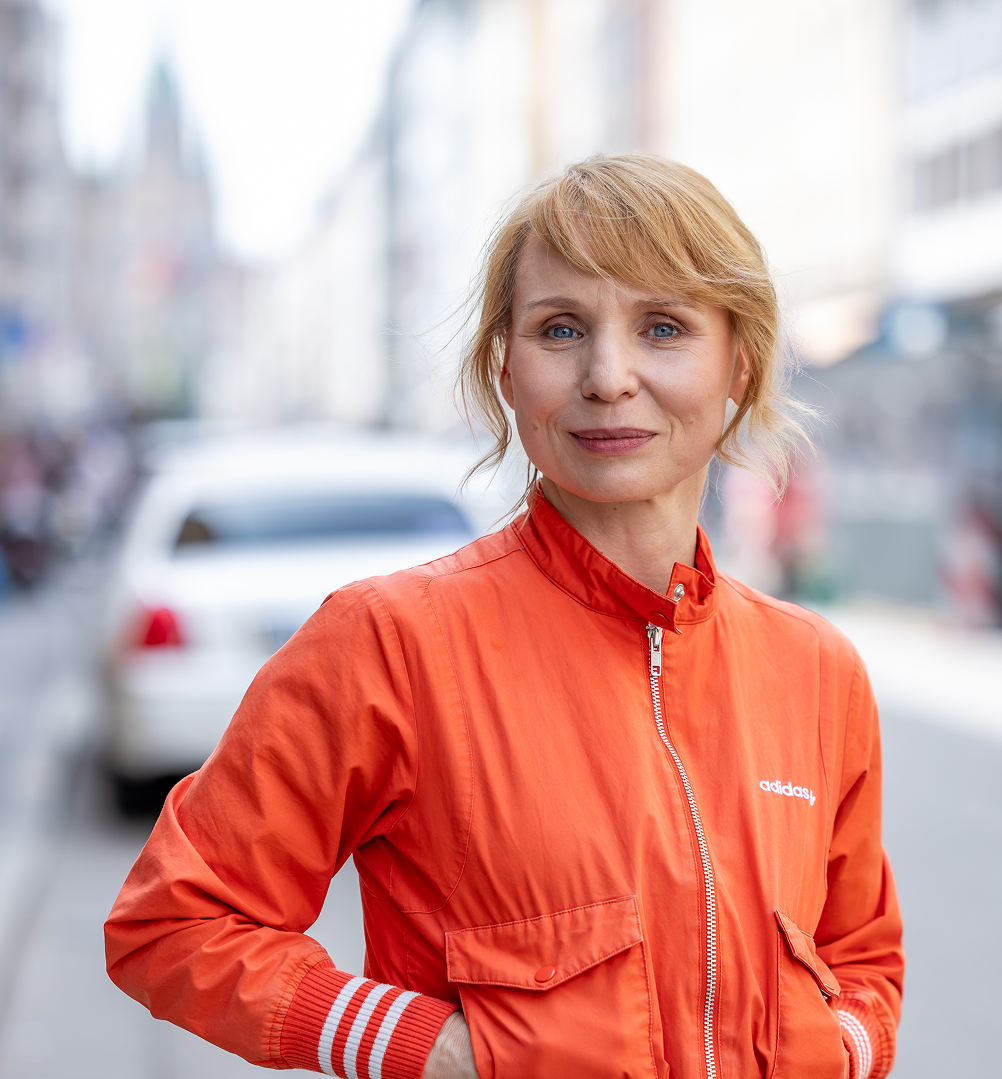 Blonde woman with blue eyes wearing an orange Adidas jacket standing on a city street.