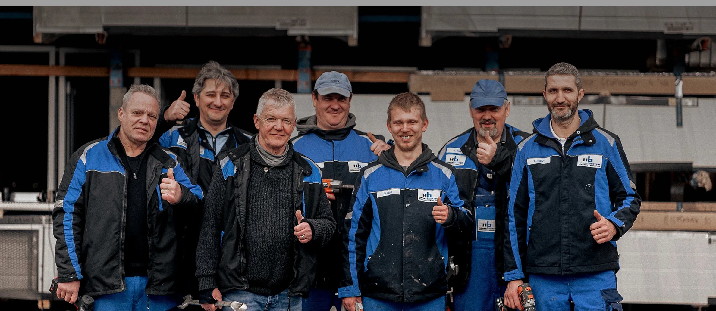 Sieben Handwerker in blauen Arbeitsjacken stehen nebeneinander in einer Werkstatt und zeigen mit Daumen nach oben.