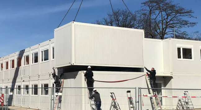 Mehrere Bauarbeiter montieren einen großen weißen Containerblock auf einem modularen Gebäudekomplex mit Leitern bei klarem Himmel.