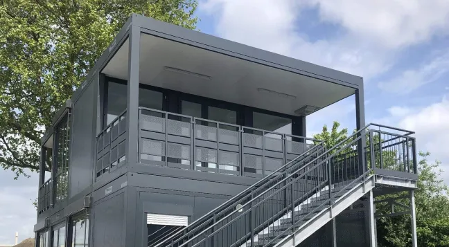 Modern zweistöckiges Gebäude aus grauen Metallcontainern mit großer Außentreppe und Geländer, umgeben von Bäumen unter blauem Himmel.