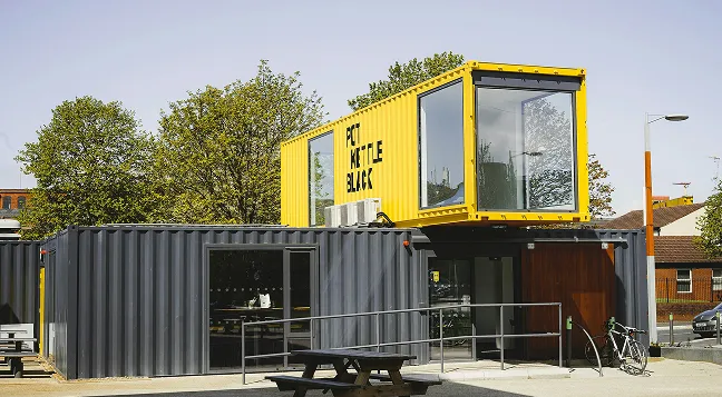 Gebäude aus schwarzen und gelben Schiffscontainern mit breiten Fenstern und einer Sitzbank davor.