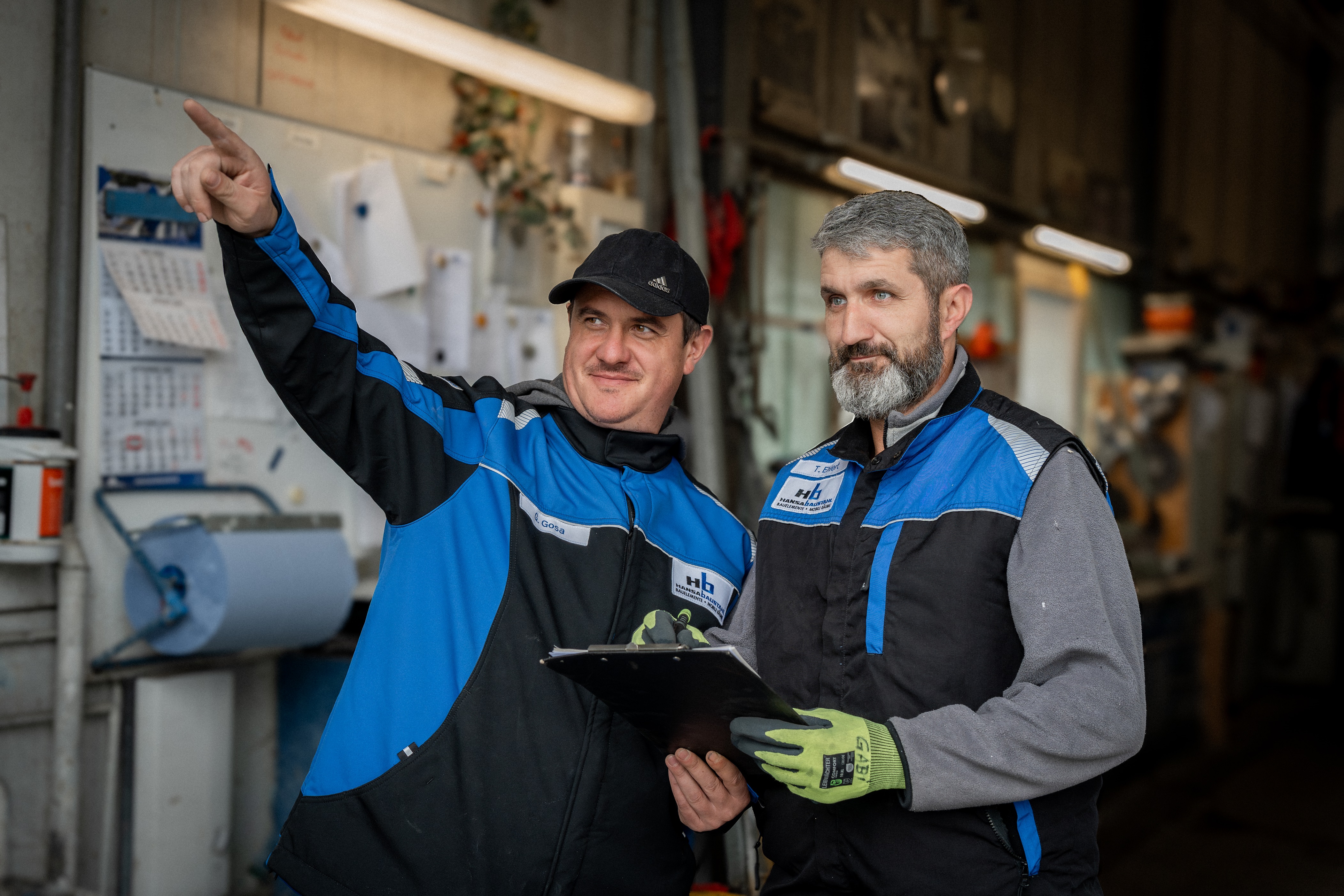Zwei Männer in blauen Arbeitsjacken besprechen sich in einer Werkstatt, einer zeigt mit dem Finger nach links, der andere hält ein Klemmbrett.