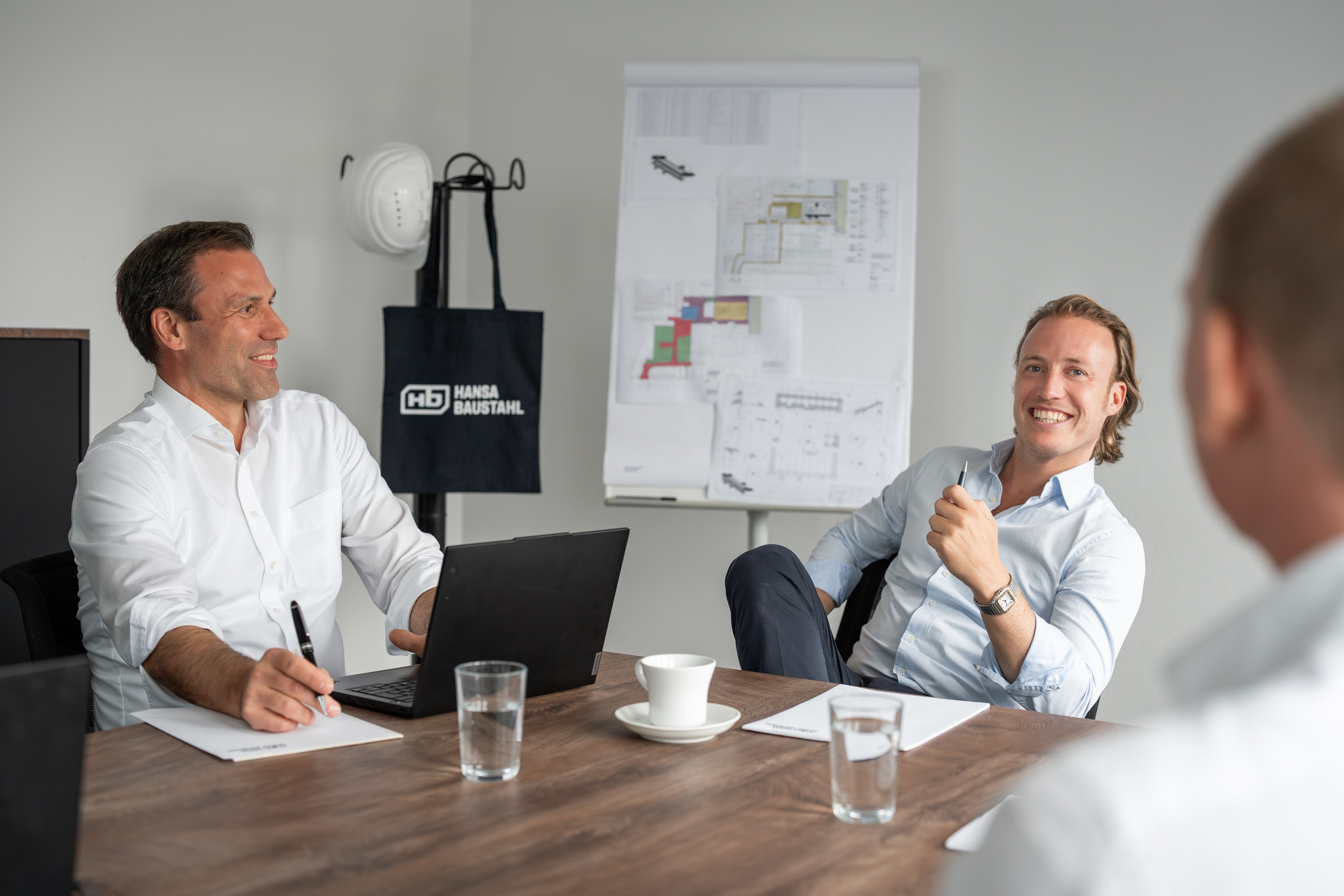 Drei Männer in einem Meetingraum, zwei lächeln und sitzen an einem Holztisch mit Laptop, Notizblöcken und Gläsern Wasser.