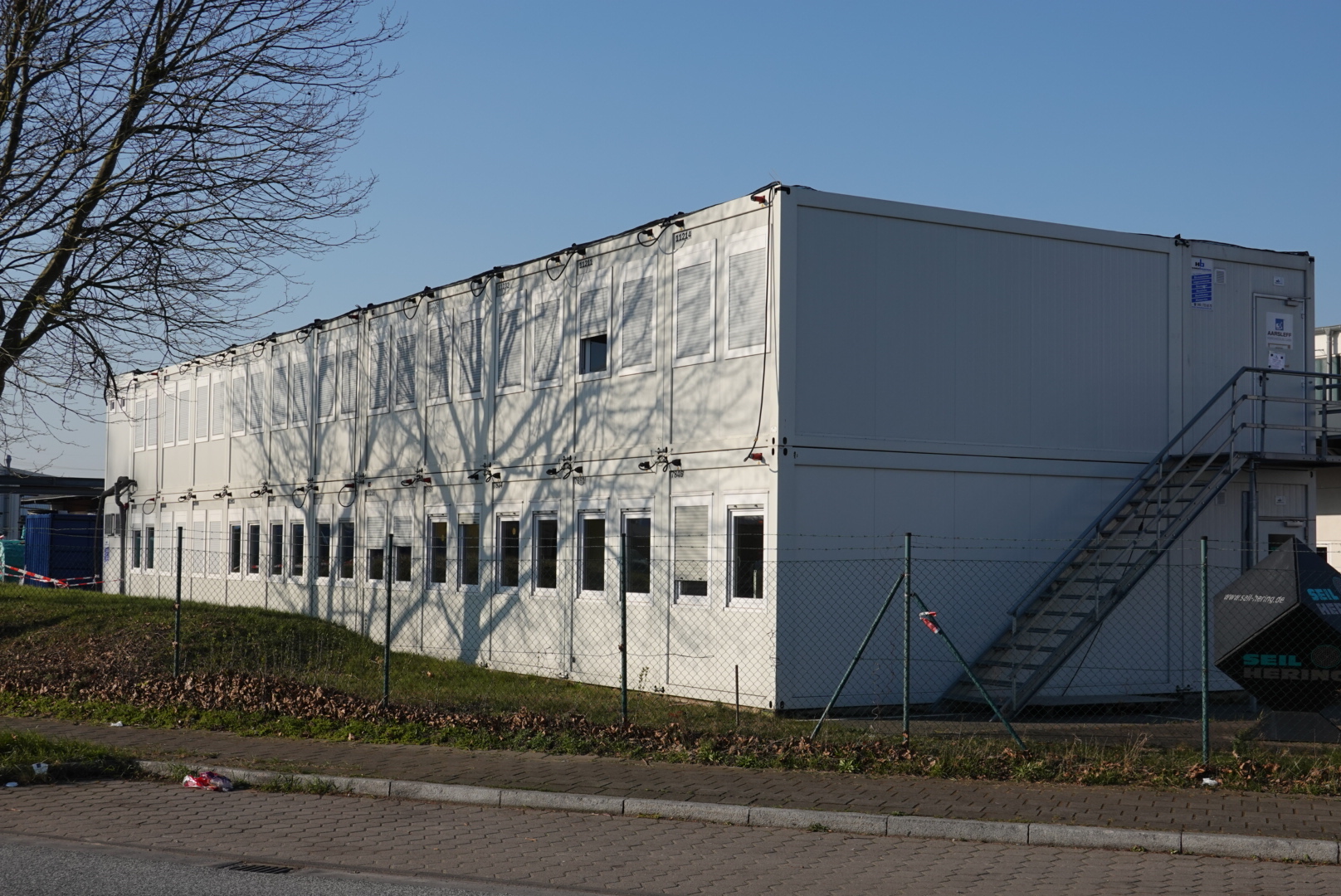 Zweistöckige weiße Baucontaineranlage mit vielen Fenstern, umgeben von einem Metallgitterzaun, bei klarem Himmel.