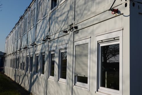 Mehrere weiße, gestapelte Modulcontainer mit Fenstern und Rolläden, von Baumzweigen beschattet.