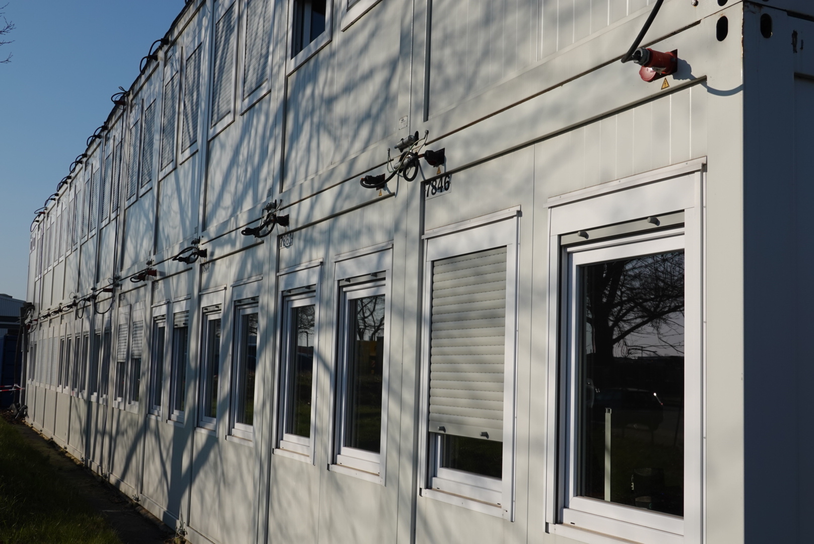 Mehrere weiße, gestapelte Modulcontainer mit Fenstern und Rolläden, von Baumzweigen beschattet.