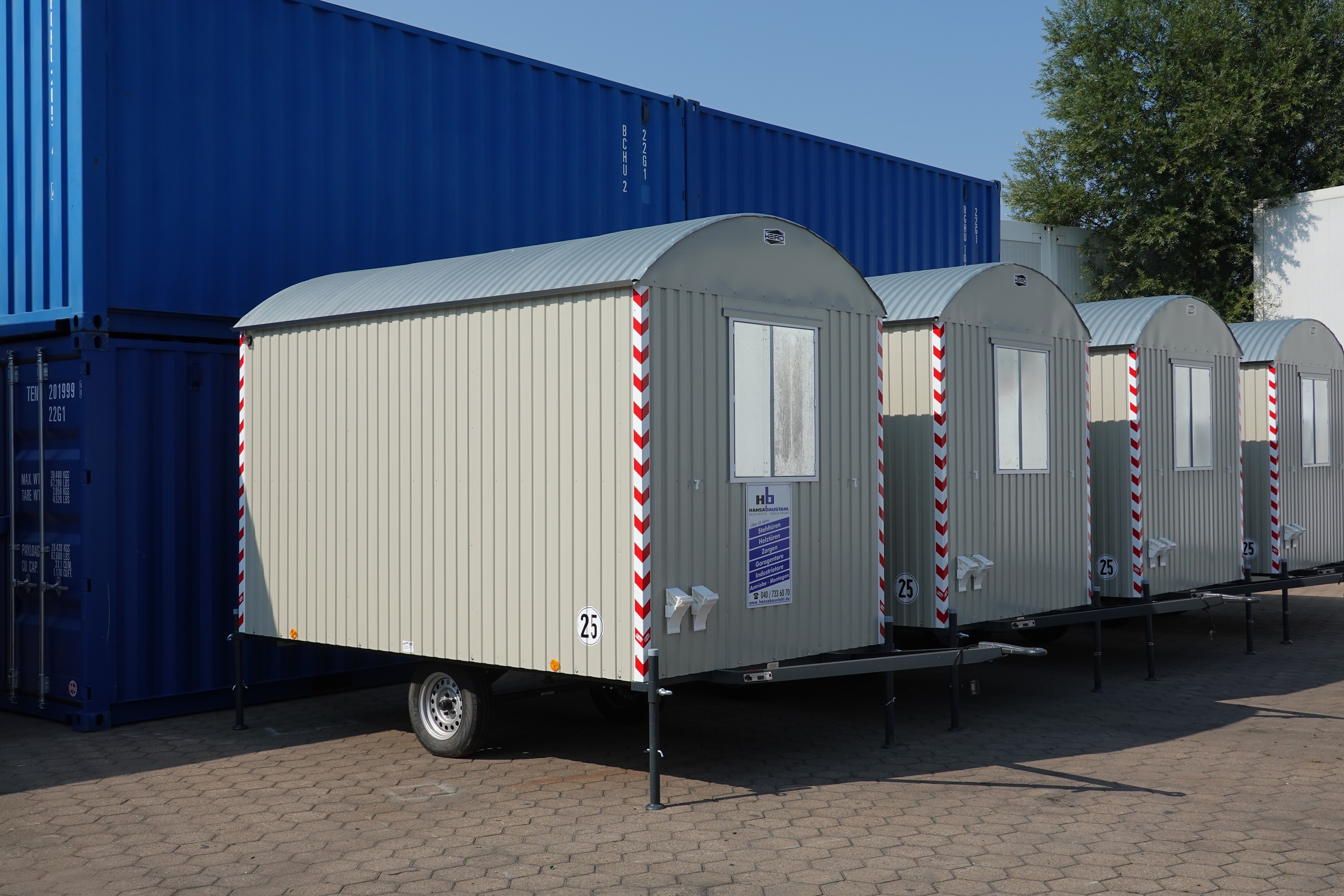 Vier mobile Bauwagen mit gewölbten, grauen Dächern und beige Seitenwänden, nebeneinander auf einem gepflasterten Hof, im Hintergrund blaue Container.