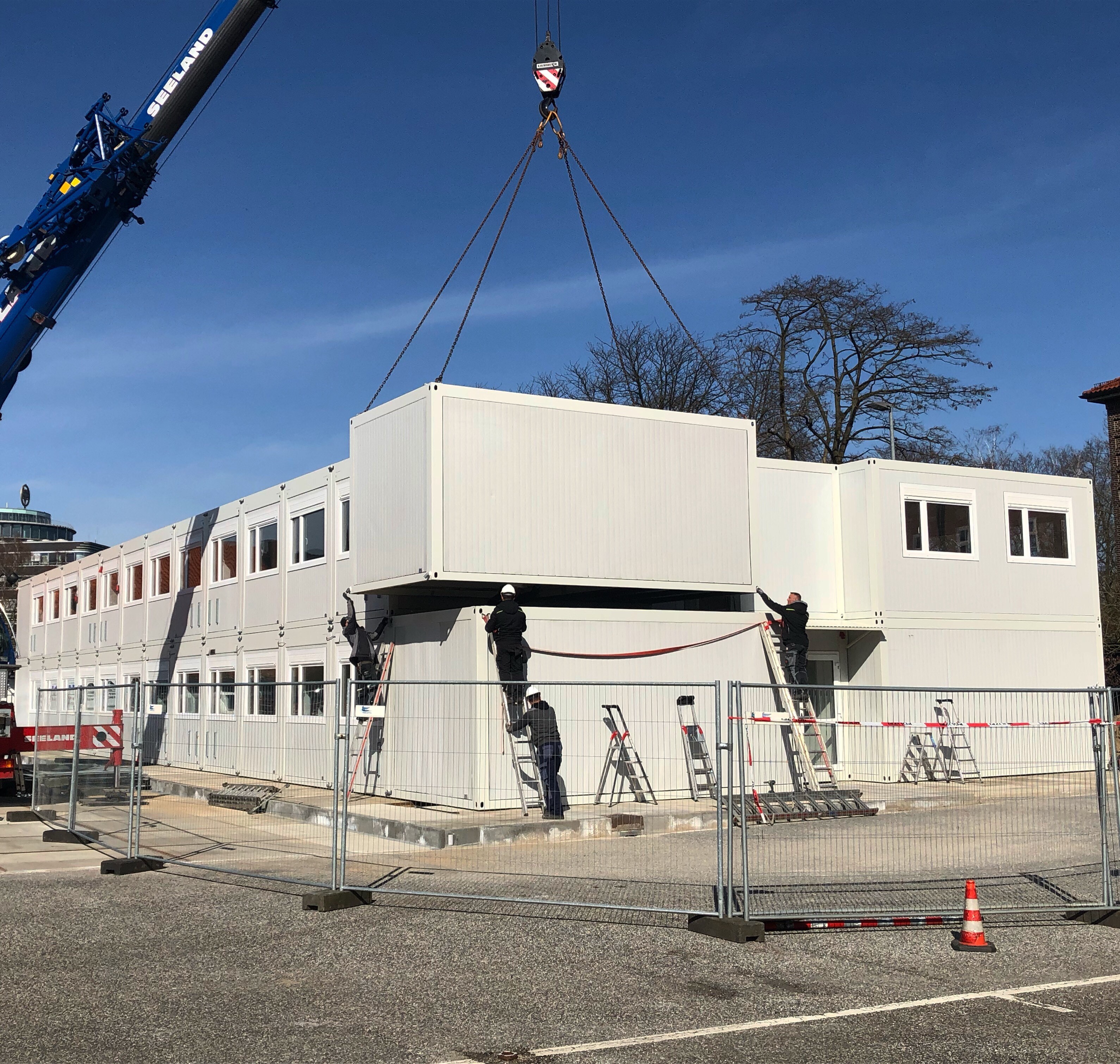 Kran hebt einen weißen Container auf eine mehrstöckige Containergebäudestruktur, während Arbeiter mit Helmen Leitern benutzen.